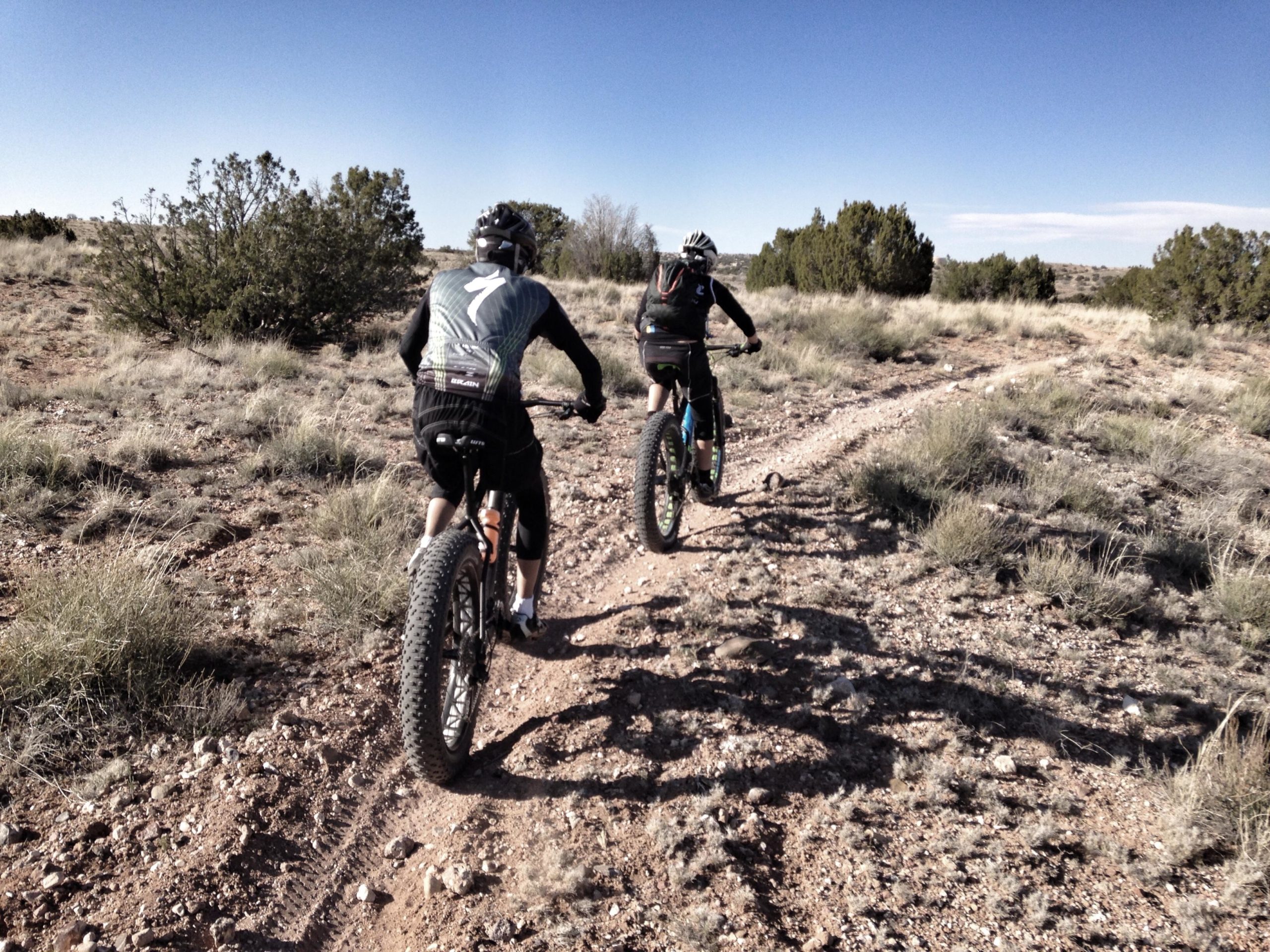 Two cyclists riding fat-tire bikes along a dirt trail in a rural landscape, surrounded by sparse vegetation and under a clear blue sky. The scene captures the adventure of mountain biking in an open natural environment. Mariposa Fat Bike Trails mountain bike trail.