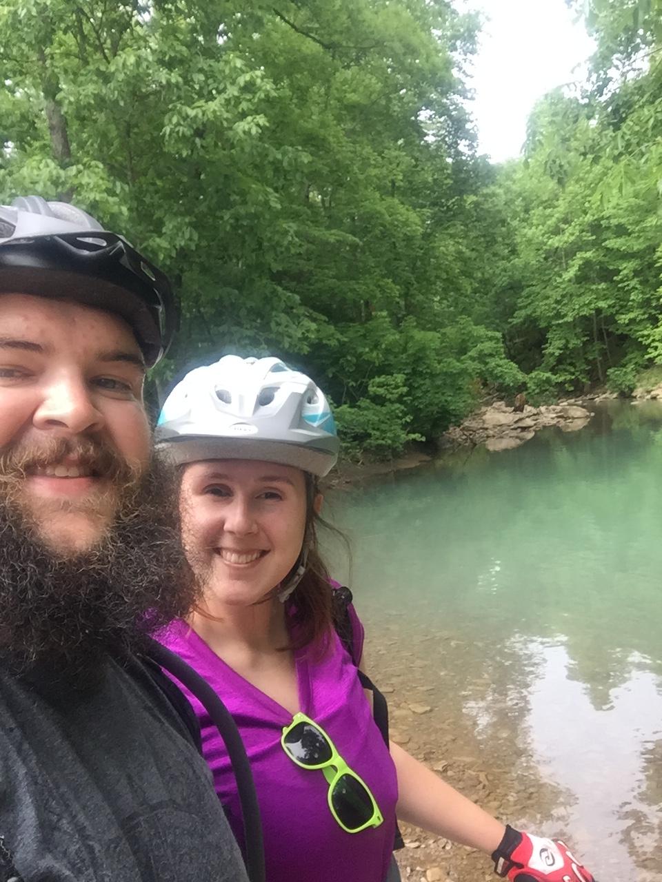 A smiling couple posing for a selfie by a serene, turquoise body of water surrounded by lush green trees, both wearing helmets and casual outdoor clothing. The woman is wearing neon green sunglasses, and the man has a dark beard. The setting suggests a fun and adventurous day spent biking or exploring nature. Fossil Flats mountain bike trail.