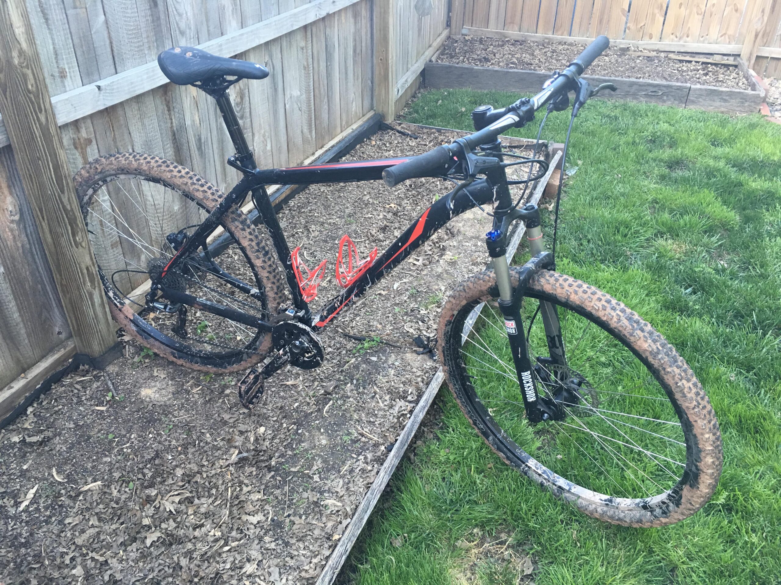 Specialized Hardrock Sport Disc 29er: A black mountain bike with orange accents is propped up against a wooden fence, featuring muddy tires and a dirty frame, indicating recent use. The bike is parked on grass and surrounded by a small garden area with wooden borders.