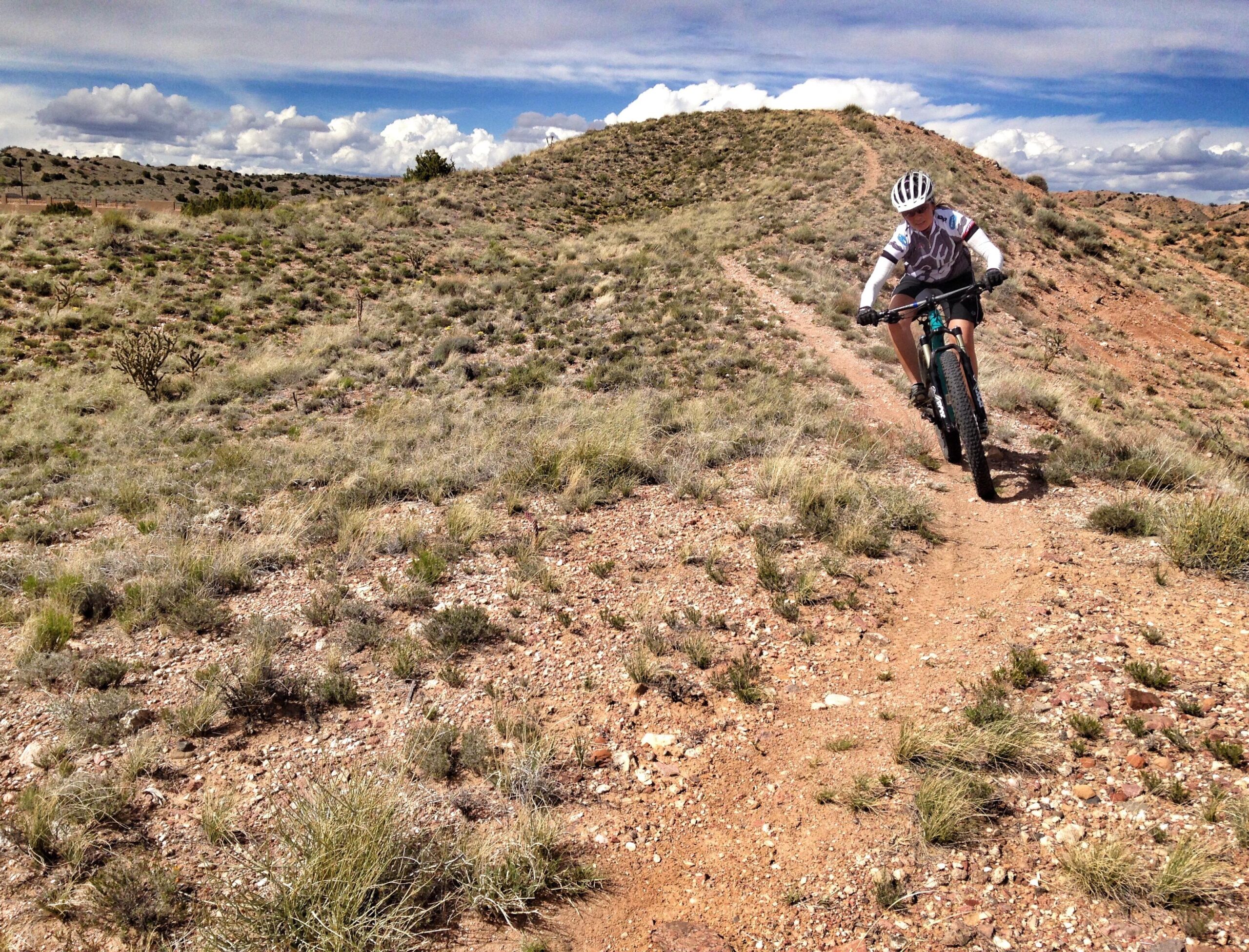 A mountain biker navigates a dirt trail on a rocky hillside, surrounded by sparse vegetation and under a partly cloudy sky. The landscape features rolling hills and a rugged terrain, showcasing a vibrant outdoor setting. Mariposa Fat Bike Trails mountain bike trail.