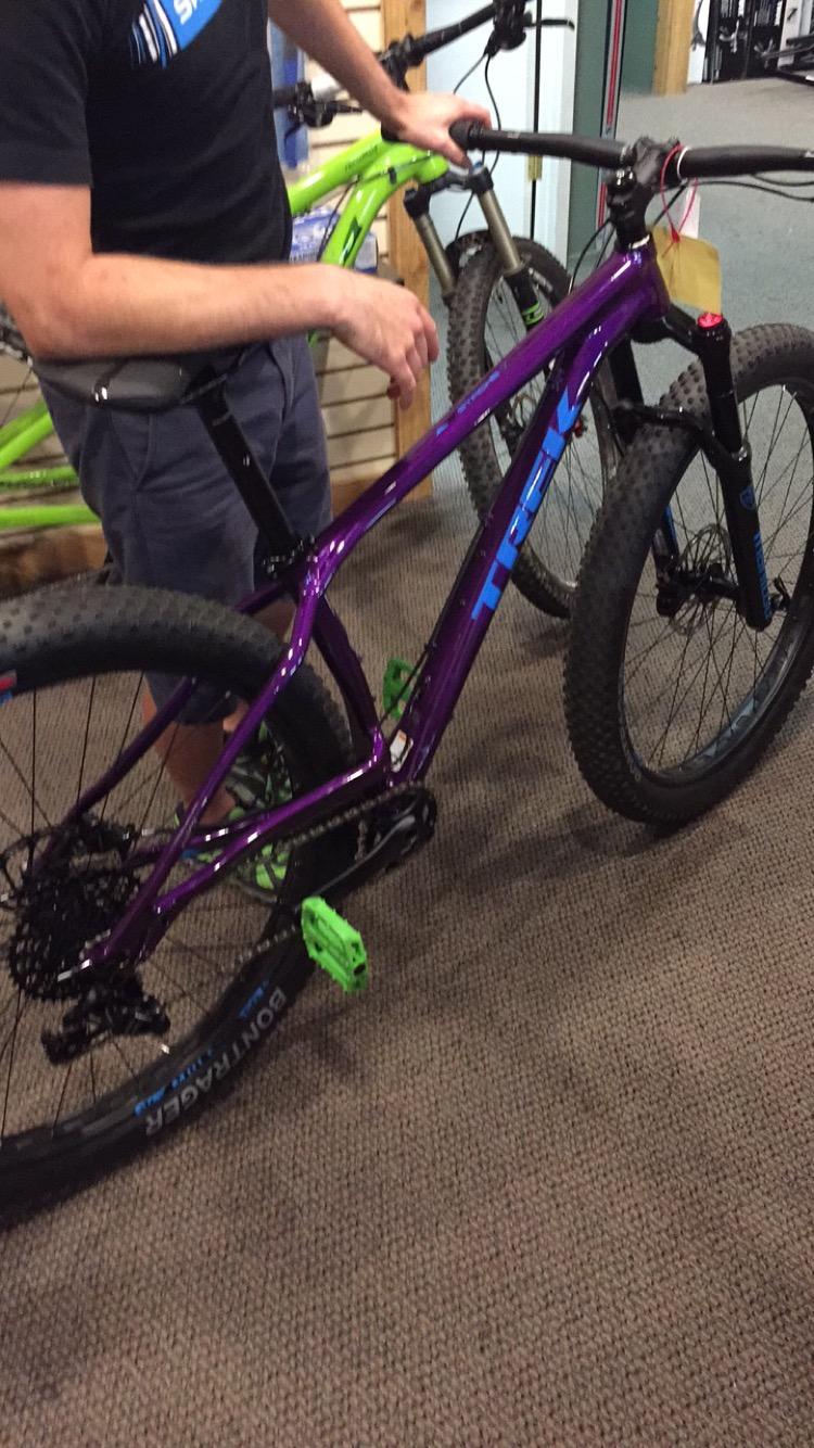 Trek Stache: A person examining a vibrant purple mountain bike with green pedals and blue accents in a bike shop, with another bike visible in the background. The flooring is carpeted, and the shop has wooden paneling on the walls.