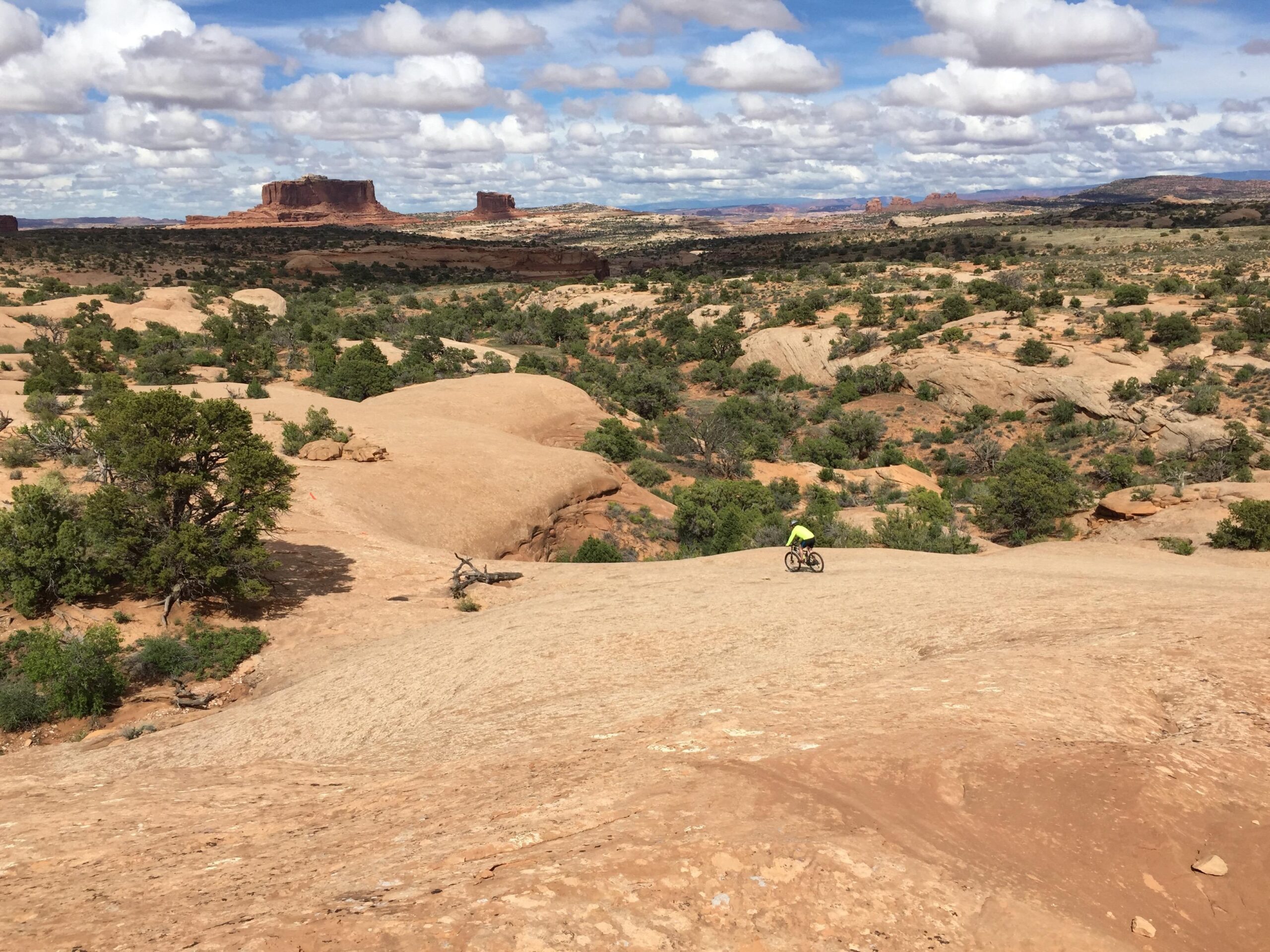 A mountain biker wearing a bright yellow shirt rides across a rocky terrain dotted with vegetation. In the background, large rock formations and a vast landscape stretch under a partly cloudy blue sky. Navajo Rocks mountain bike trail.