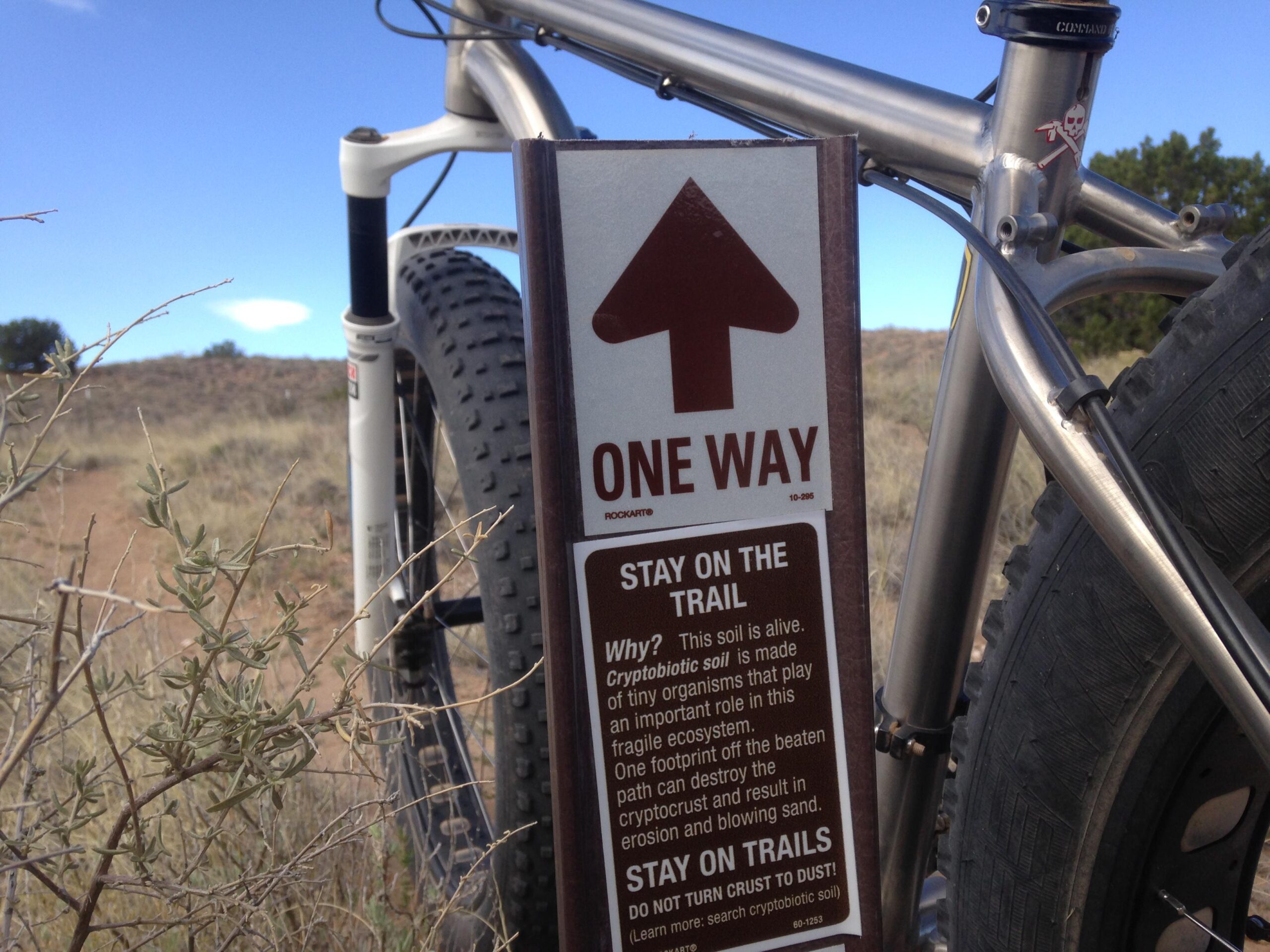 A close-up of a "One Way" trail sign with a bicycle in the background, set against a dry, natural landscape. The sign reads "Stay on the Trail," providing information about cryptobiotic soil and its importance in the ecosystem, with an upward arrow indicating the direction of travel. The scene features sparse vegetation and a blue sky. Mariposa Fat Bike Trails mountain bike trail.
