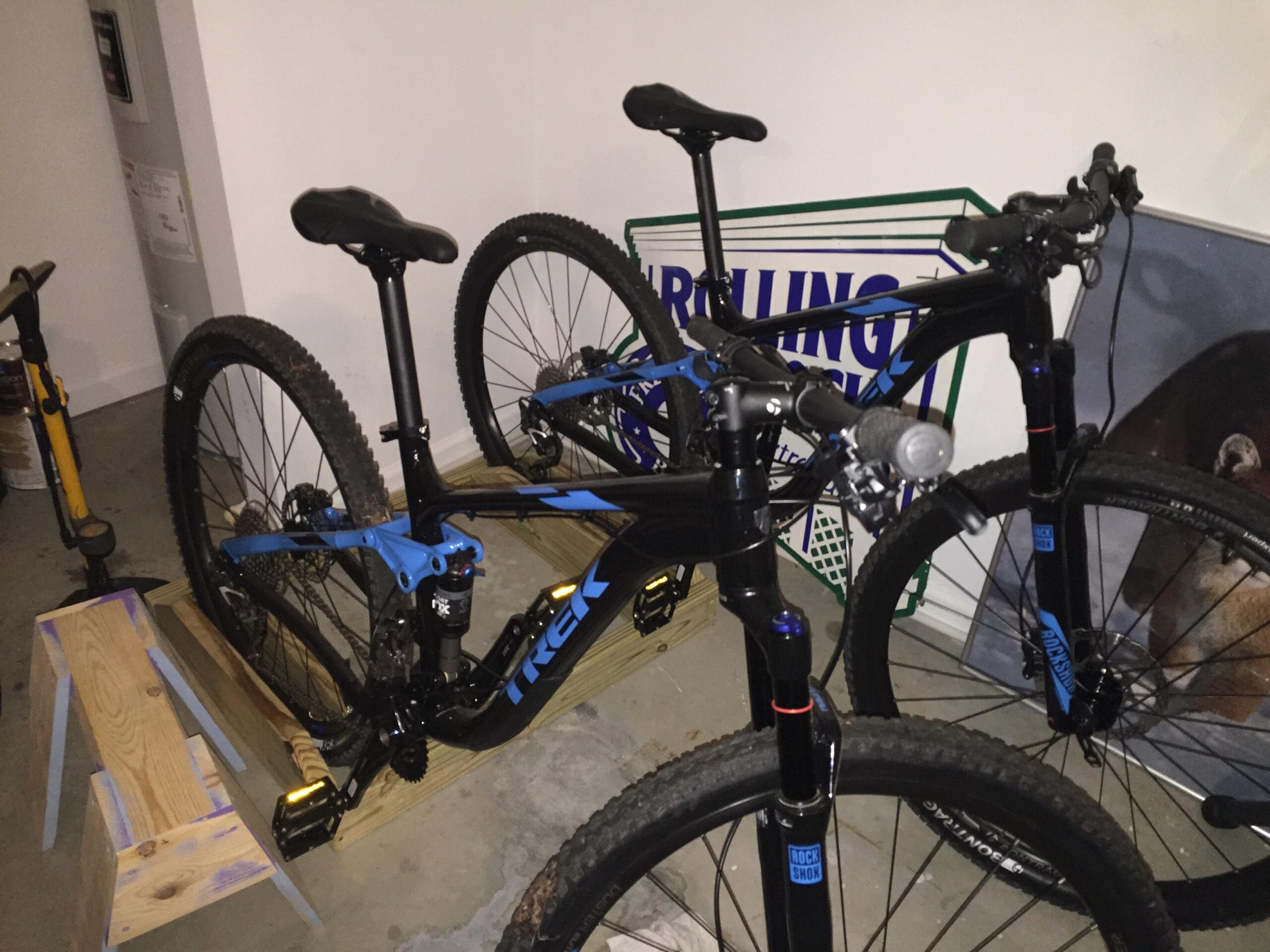 Trek Fuel EX 7 29: Two mountain bikes with black and blue frames are parked in a garage. One bike is positioned on a wooden stand, while the other stands next to it. The background includes a partially visible poster and a yellow air pump. The garage floor is concrete, and there are various items in the surrounding space.