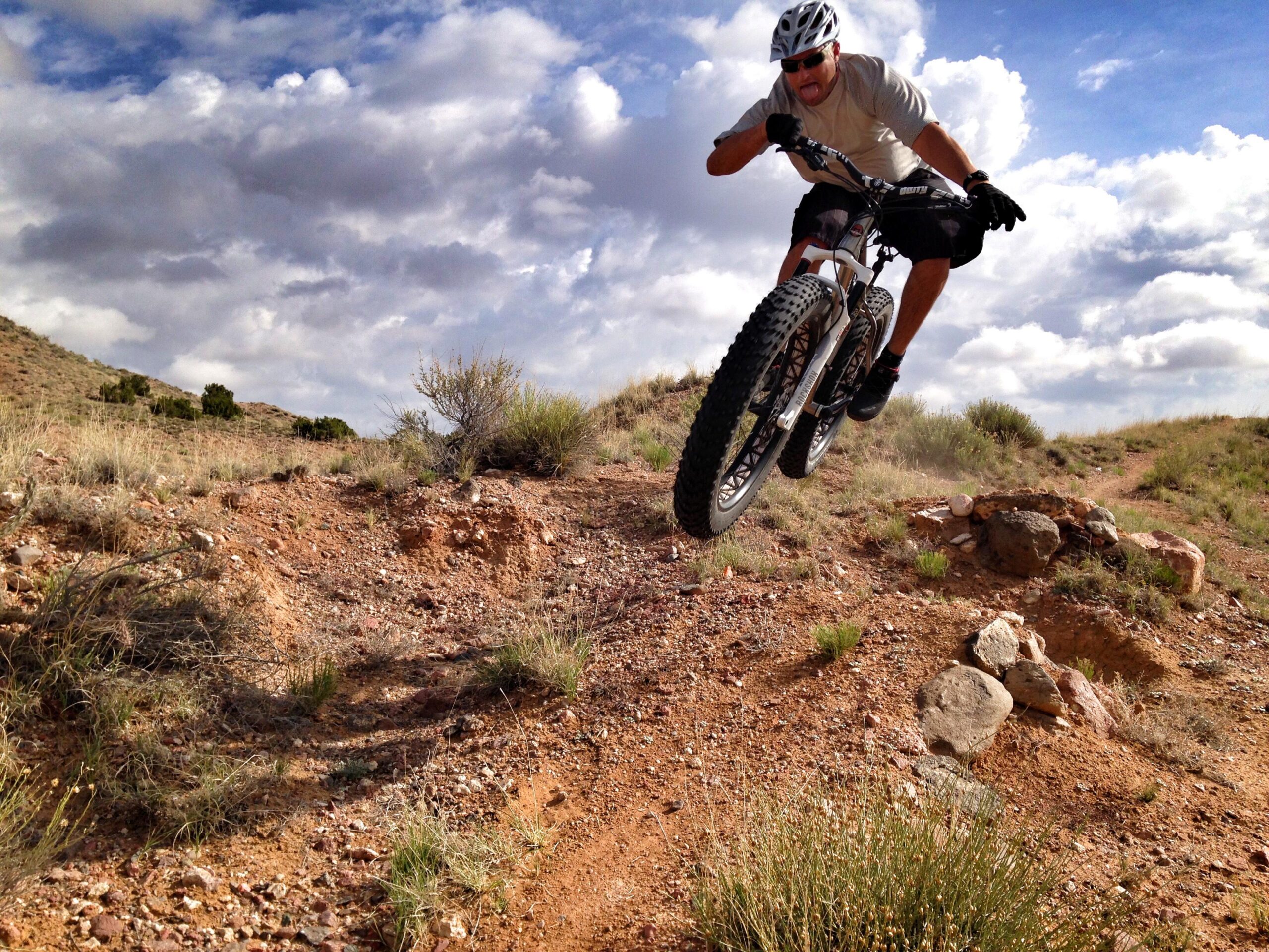 A mountain biker performing a jump over rough terrain with dust trailing behind. The background features a hilly landscape with sparse vegetation and a partly cloudy sky. The cyclist is wearing a helmet and protective gear, showcasing an action-packed moment in outdoor cycling. Mariposa Fat Bike Trails mountain bike trail.
