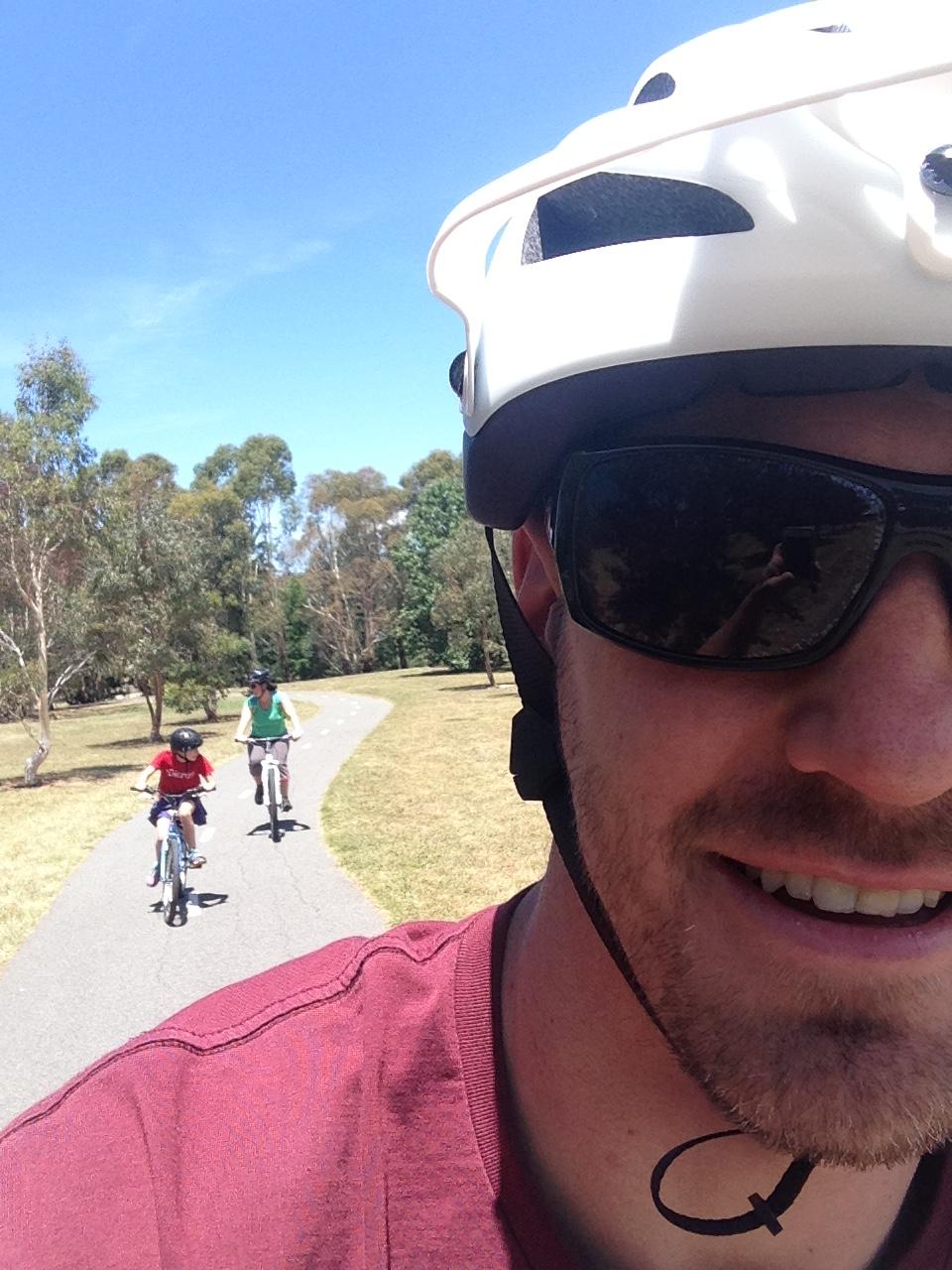 Giant Anthem X 29er 1: A person wearing sunglasses and a white bicycle helmet smiles at the camera while riding a bike on a sunny path. In the background, two children, one in a red shirt and the other in a green shirt, are also riding their bikes along the same path, surrounded by trees and grass.