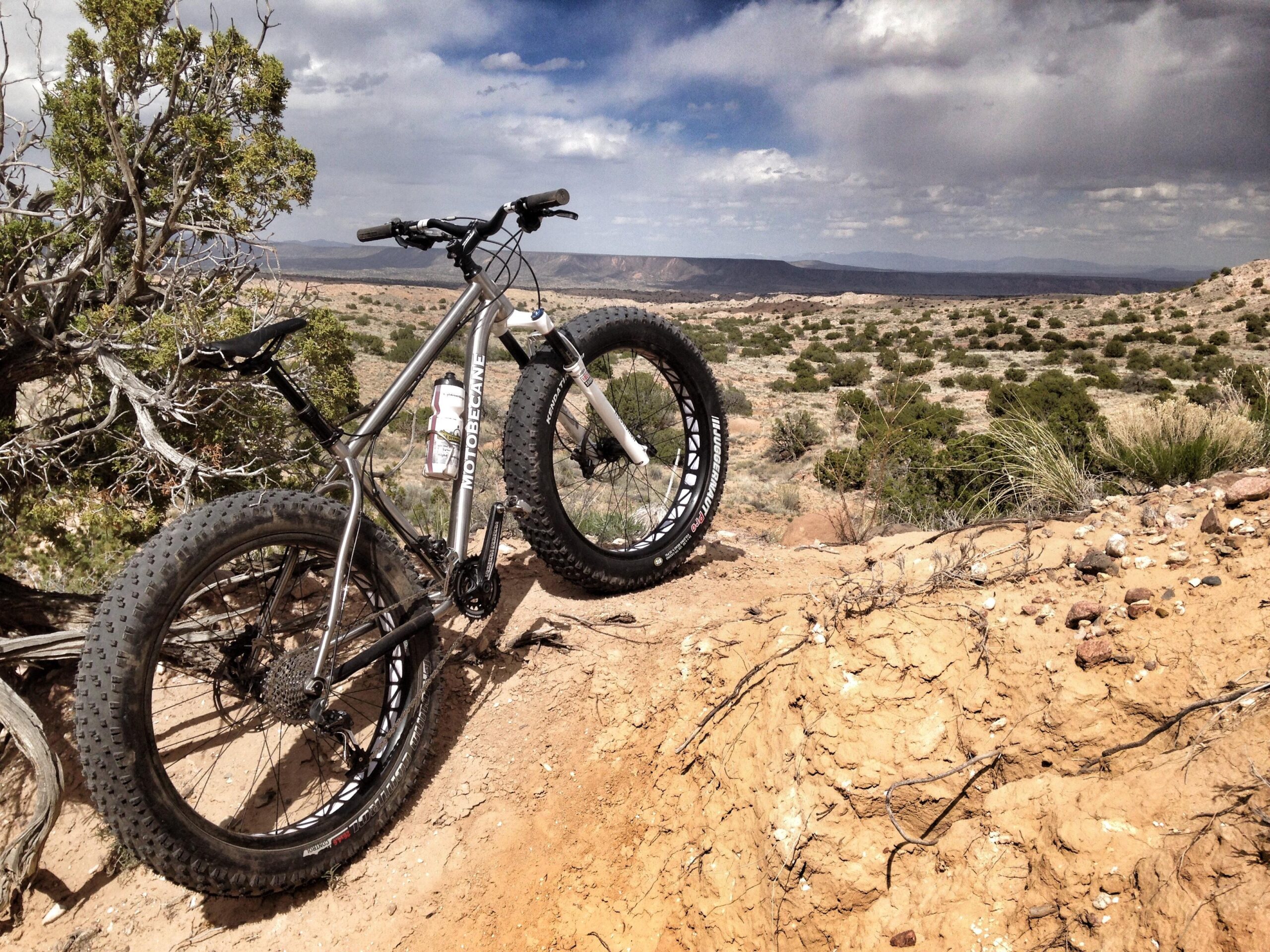 A fat tire mountain bike resting on a rocky, sandy hill overlooking a vast desert landscape with sparse vegetation and distant mountains under a partly cloudy sky. Mariposa Fat Bike Trails mountain bike trail.