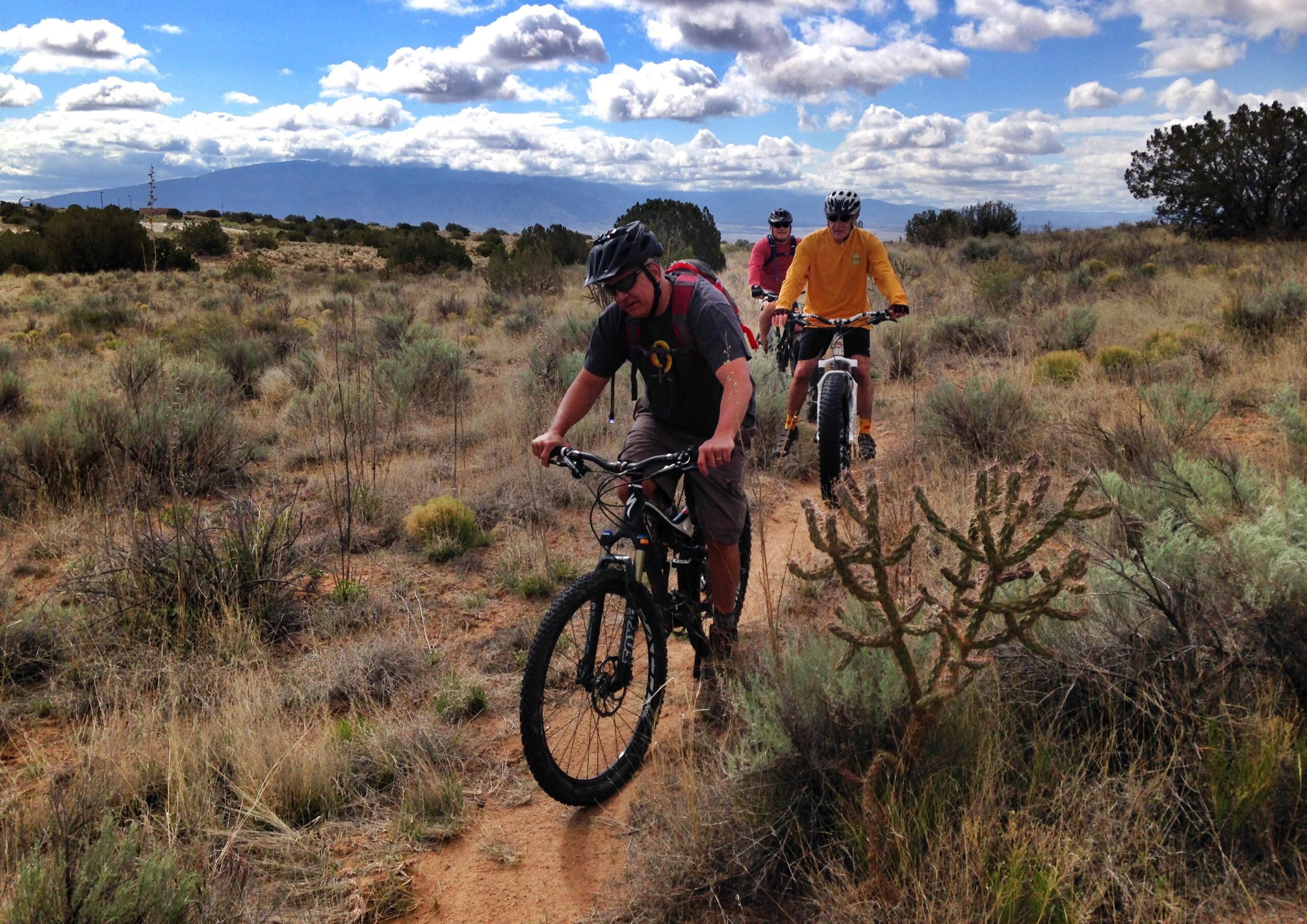 Three mountain bikers navigate a dirt trail surrounded by desert vegetation and low shrubs. In the background, there are rolling hills under a partly cloudy sky. The scene captures the essence of outdoor adventure and cycling in a natural landscape. Mariposa Fat Bike Trails mountain bike trail.