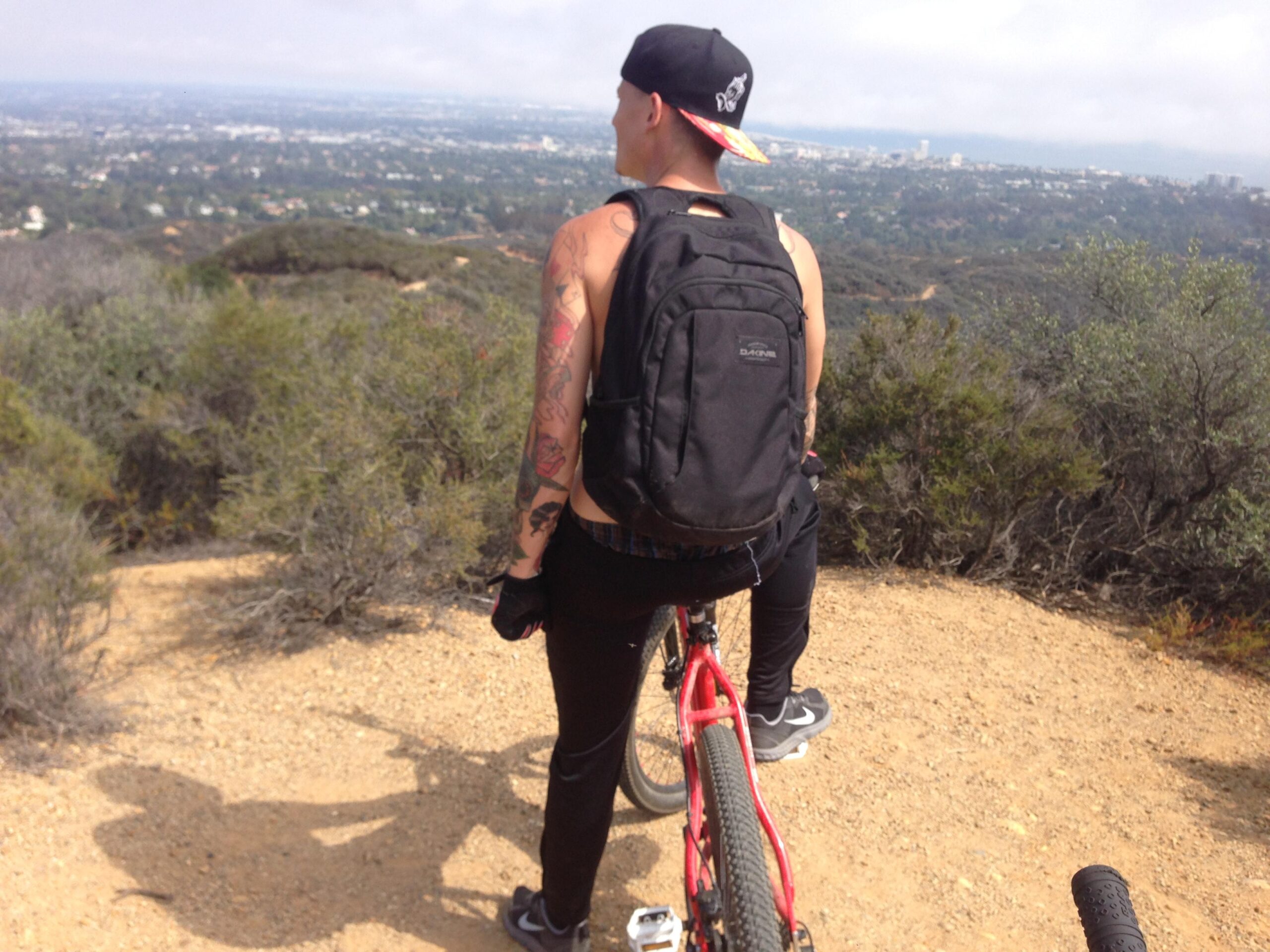 Haro Flightline Sport: A person with tattoos, wearing a cap and a backpack, is standing beside a red mountain bike on a dirt trail. They are looking out over a scenic view of a city landscape and distant hills, surrounded by sparse vegetation and shrubs. The sky is partly cloudy.