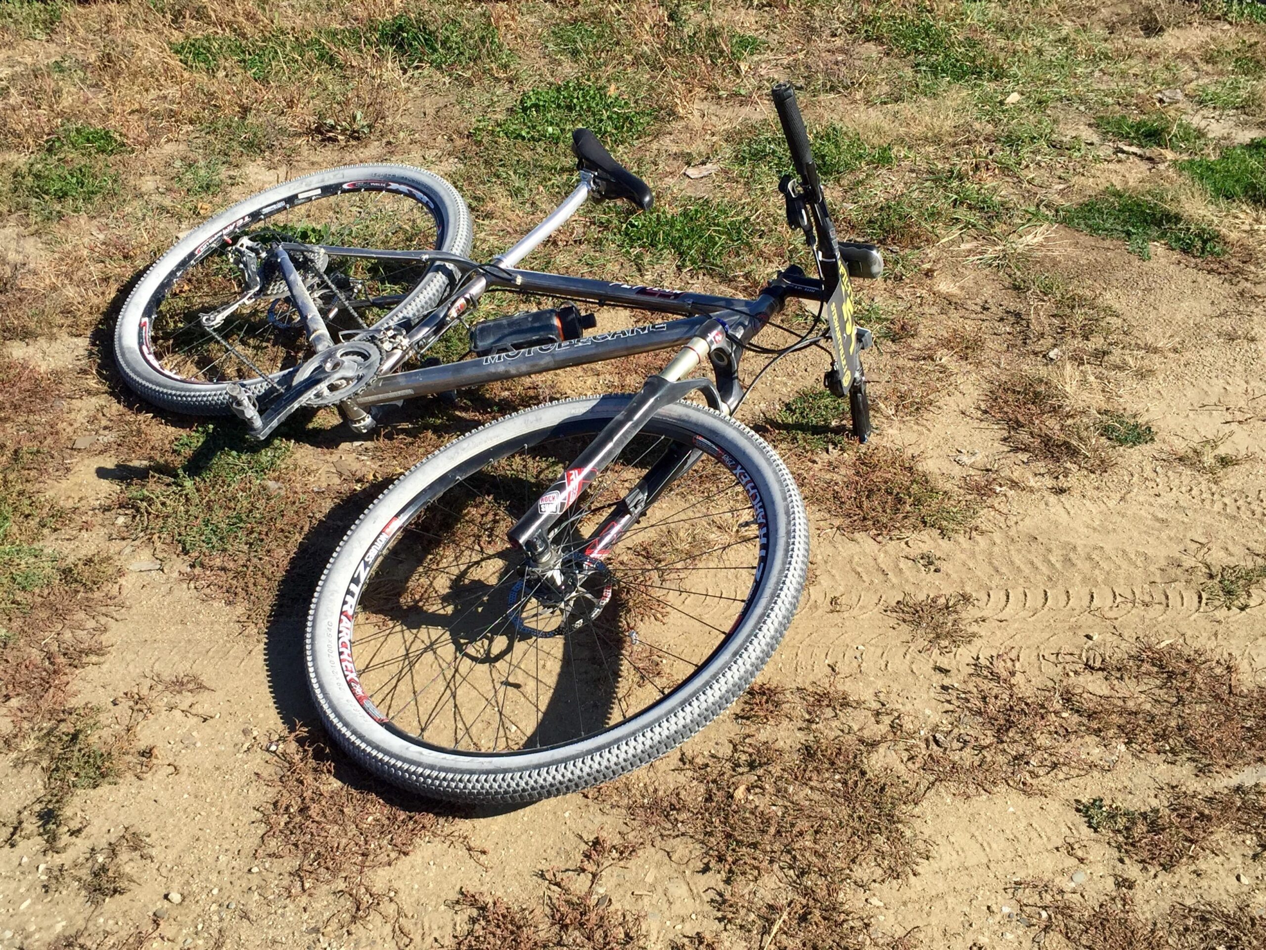 Motobecane Fly Team 29er Ti X0 Edition: A silver mountain bike lying on its side on a dirt surface, surrounded by patches of grass. The front wheel is flat, and tire tracks are visible in the dirt nearby.