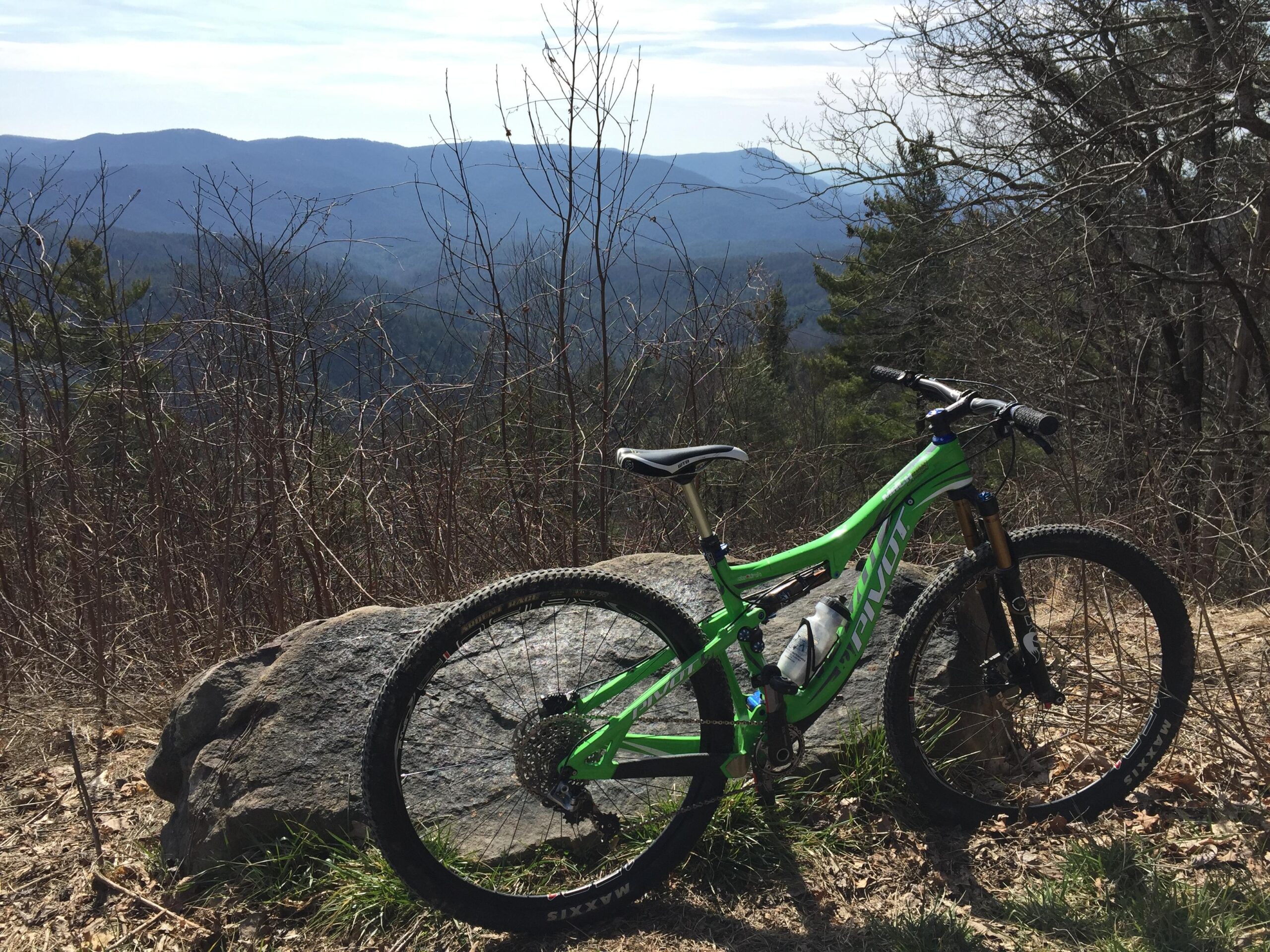 Pivot Mach 429 Carbon: A bright green mountain bike resting against a large rock, with a scenic view of rolling mountains in the background. The landscape features sparse trees and a clear sky, suggesting a sunny day.