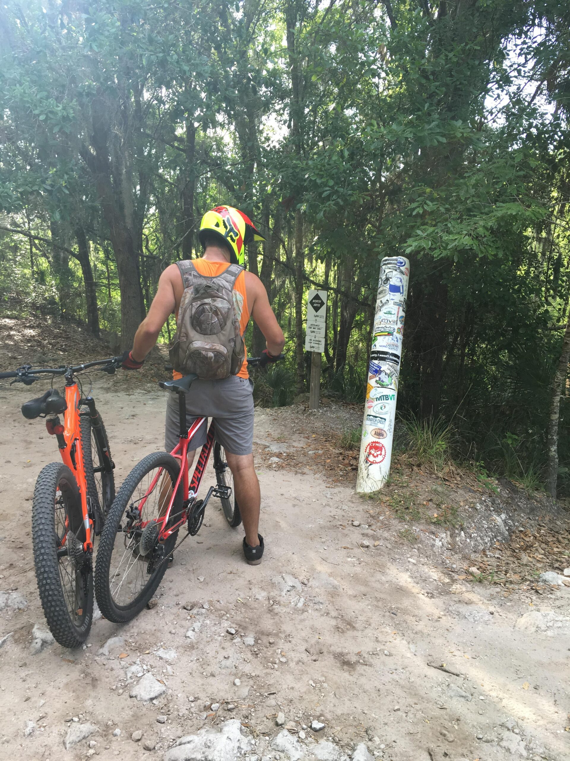 A person wearing a bright helmet and a backpack is stopped on a dirt path, holding two mountain bikes. They are facing a trail sign surrounded by lush greenery, indicating a biking route. The setting suggests an outdoor recreational area, ideal for biking. Balm Boyette Scrub Preserve mountain bike trail.