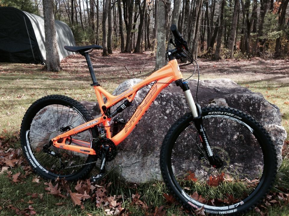 Intense tracer: A bright orange mountain bike leaning against a large rock, surrounded by a wooded area with fallen leaves. A tent is visible in the background among the trees, suggesting a camping or outdoor setting.