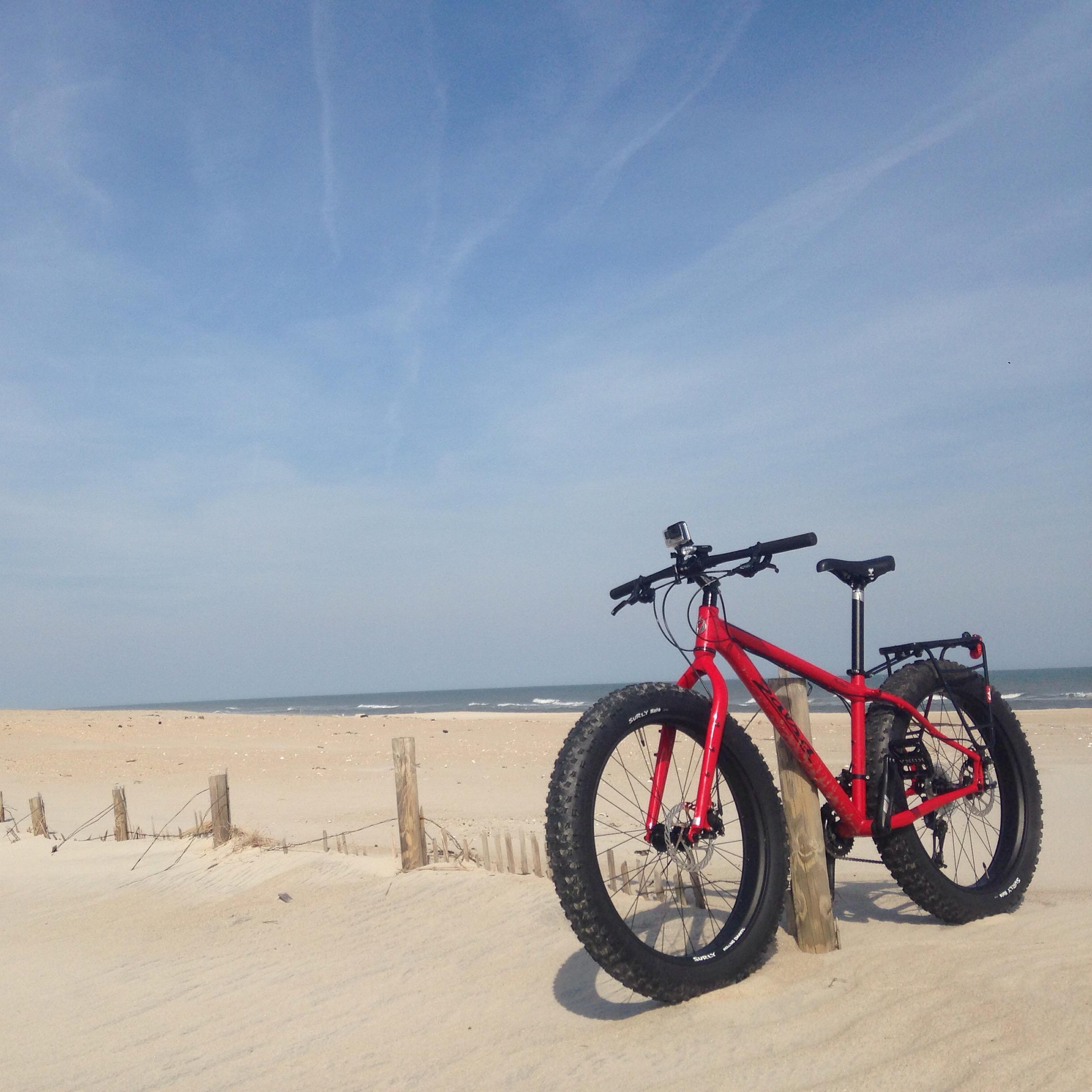 Salsa Mukluk: A red fat bike parked on a sandy beach with gentle waves in the background under a clear blue sky. Wooden fencing lines the beach, and footprints are visible in the sand.