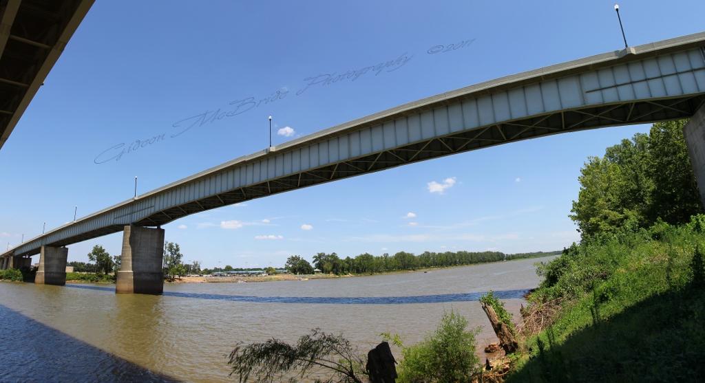 A panoramic view of a river with a bridge overhead, under a clear blue sky. The bridge features a concrete structure supported by pillars, with lush greenery lining the riverbanks. The water reflects the blue sky and clouds, creating a serene landscape. Stoner Park mountain bike trail.