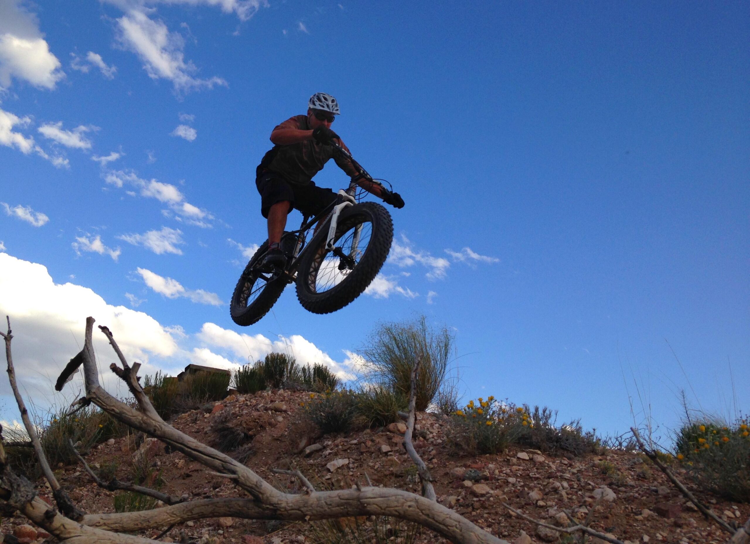 A mountain biker in mid-air, performing a jump over a rocky terrain with a blue sky and scattered clouds in the background. The biker is wearing a helmet and sunglasses, and the thick tires of the bike are clearly visible as it ascends above the ground. Some shrubbery and branches are present in the foreground. Mariposa Fat Bike Trails mountain bike trail.