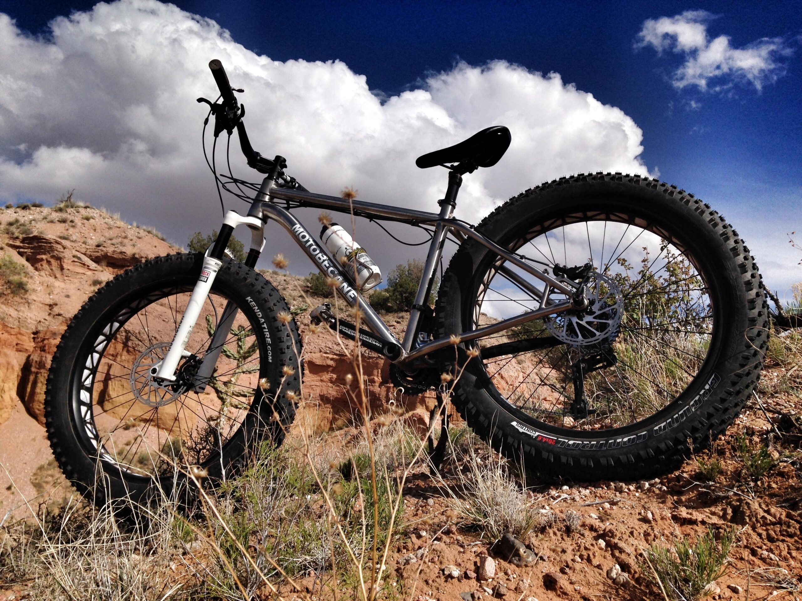 A silver mountain bike with wide tires is positioned on a sandy, rugged terrain with low vegetation. In the background, there are rocky formations and a dramatic sky filled with clouds. The image captures the bike at an angle, emphasizing its sturdy build and off-road capabilities. Mariposa Fat Bike Trails mountain bike trail.