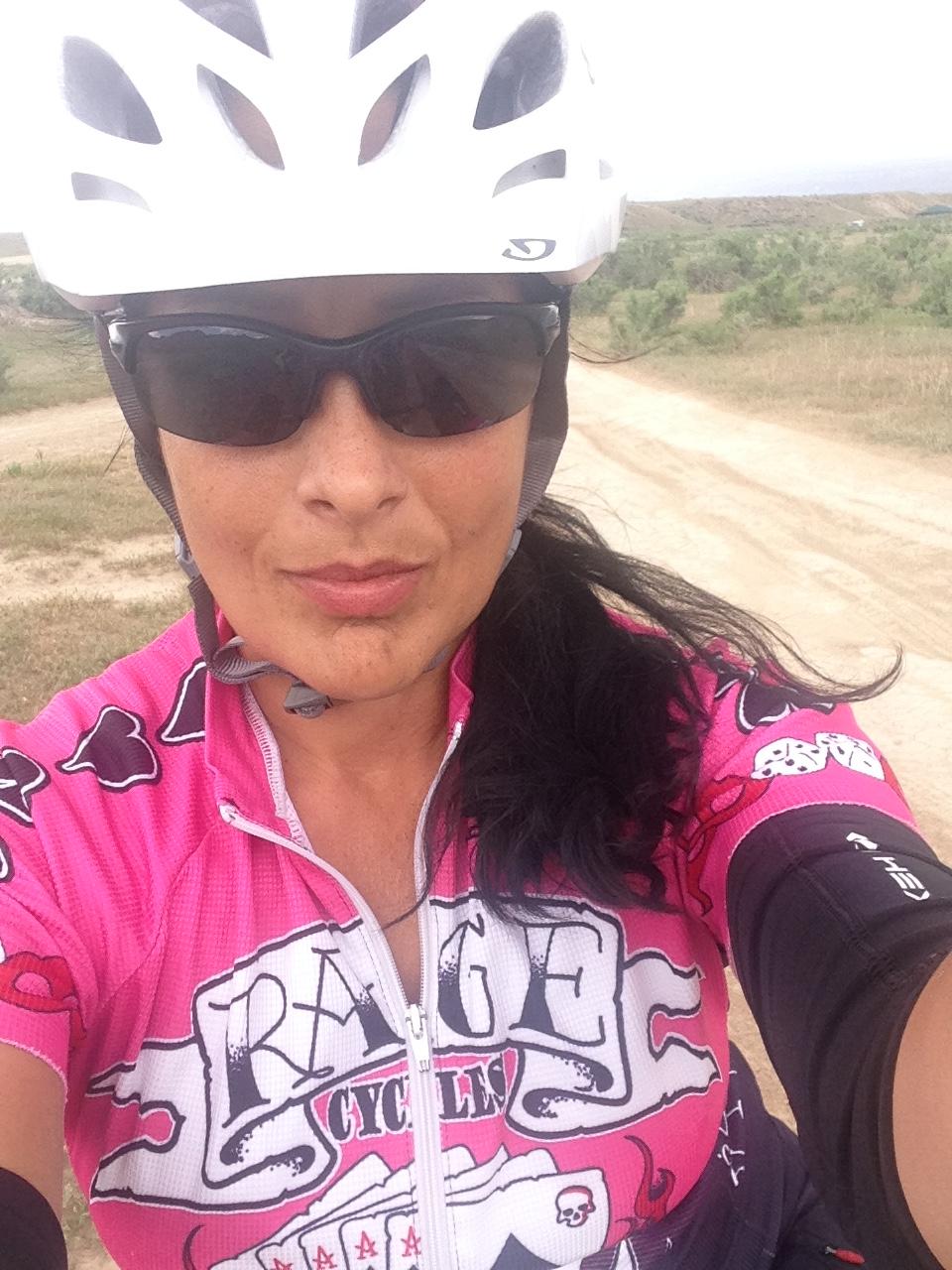 Cannondale Scalpel: Close-up selfie of a cyclist wearing a white helmet and dark sunglasses. They are dressed in a vibrant pink cycling jersey featuring a graphic design. The background shows a dirt path surrounded by greenery under a cloudy sky.