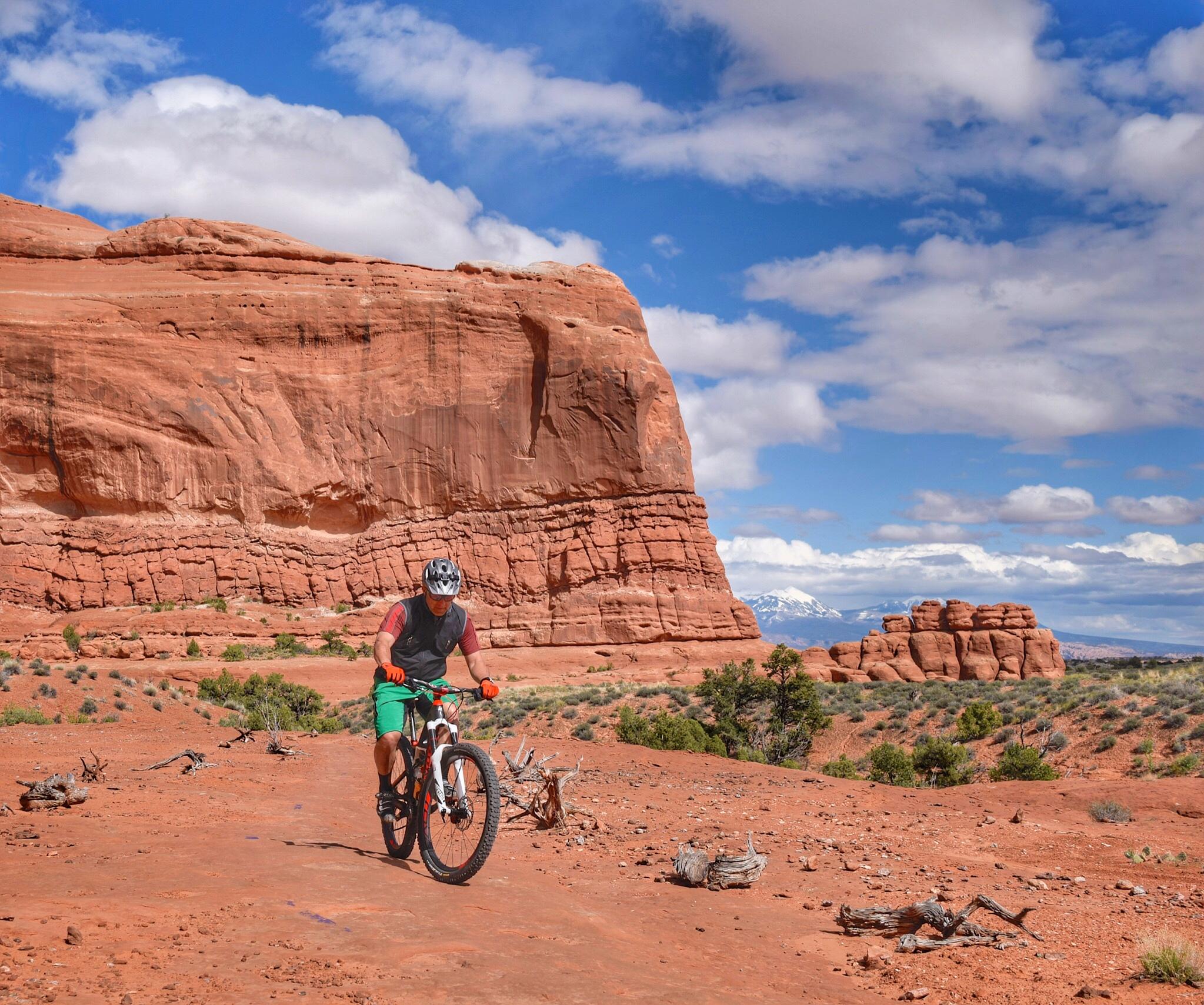A mountain biker navigating a rocky trail in a desert landscape, surrounded by red rock formations and sparse vegetation under a blue sky with fluffy clouds. Navajo Rocks mountain bike trail.
