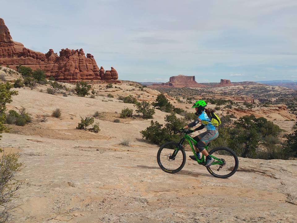 A person riding a green mountain bike on a rocky trail in a desert landscape, with distinctive red rock formations and distant plateaus in the background under a partly cloudy sky. Ramblin' mountain bike trail.