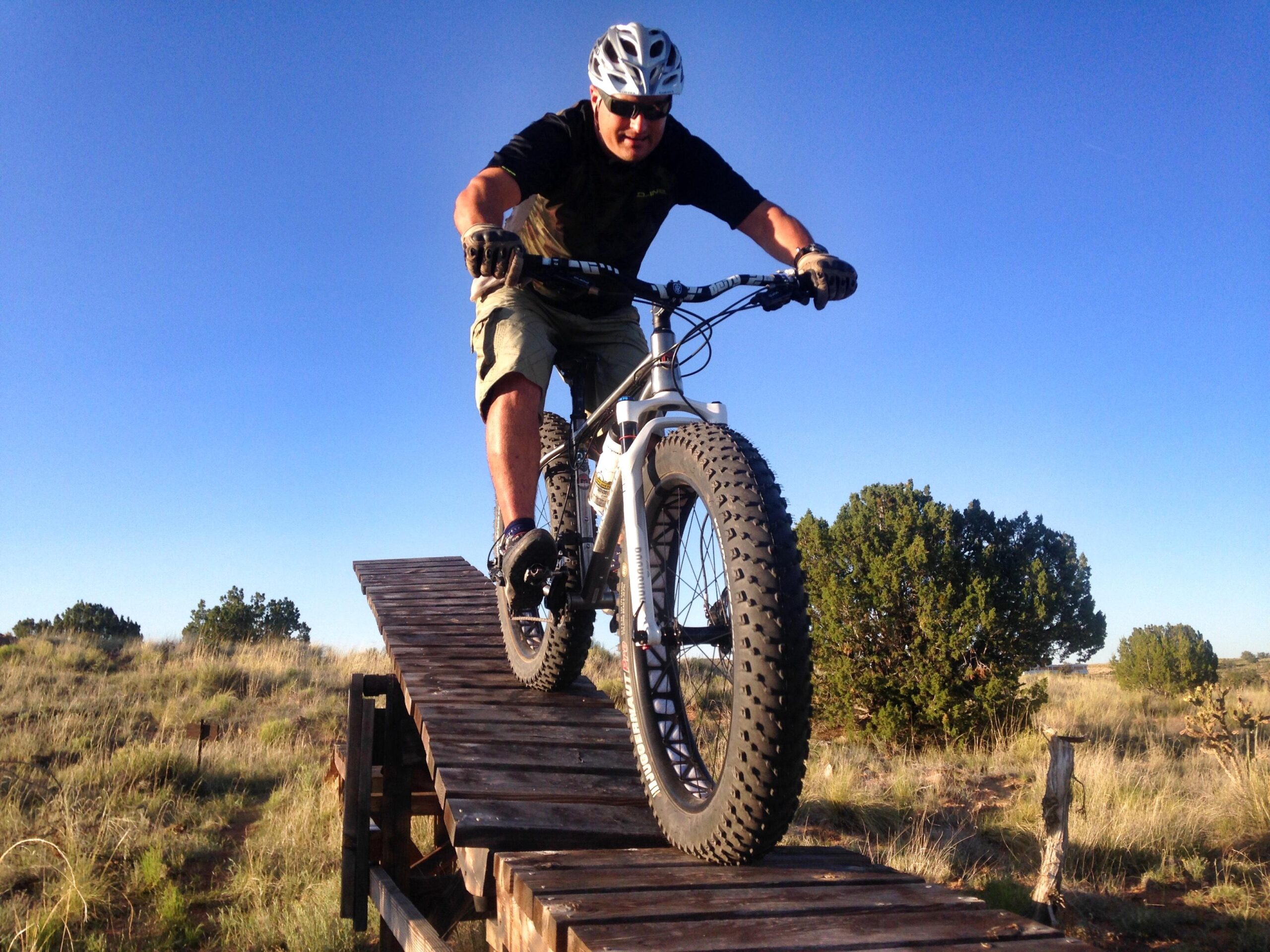 A mountain biker skillfully rides over a wooden ramp in a natural outdoor setting, with clear blue skies and sparse vegetation in the background. The biker is wearing a helmet and protective gear, showcasing an adventurous spirit.