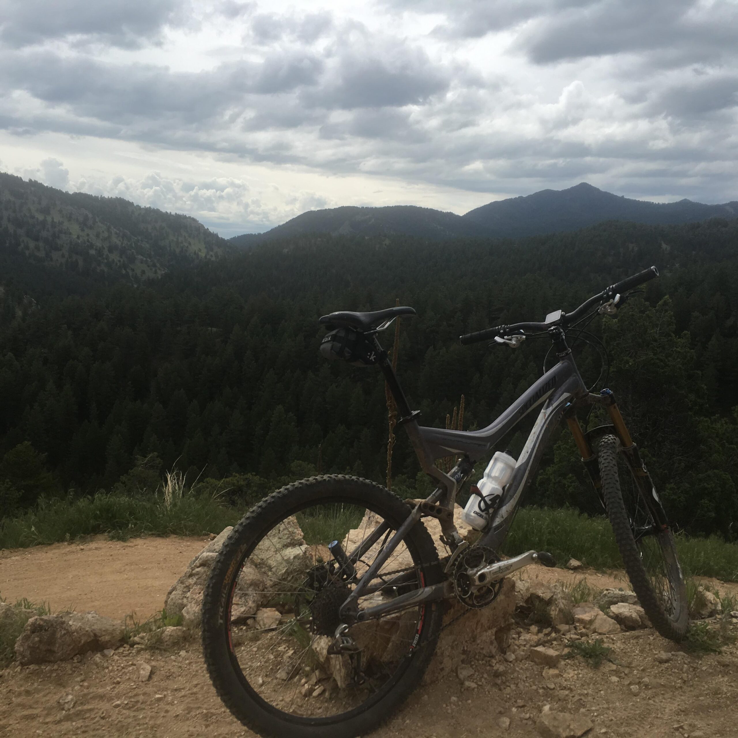 A mountain bike parked on a rocky outcrop, overlooking a lush green valley and distant mountains under a cloudy sky. Betasso Preserve mountain bike trail.