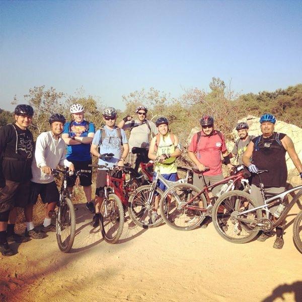 Specialized Camber Comp 29: A group of eight mountain bikers stands on a dirt path surrounded by trees and rocks. They are wearing helmets and casual athletic clothing, with different types of mountain bikes parked beside them. The sky is clear and blue, indicating a sunny day.