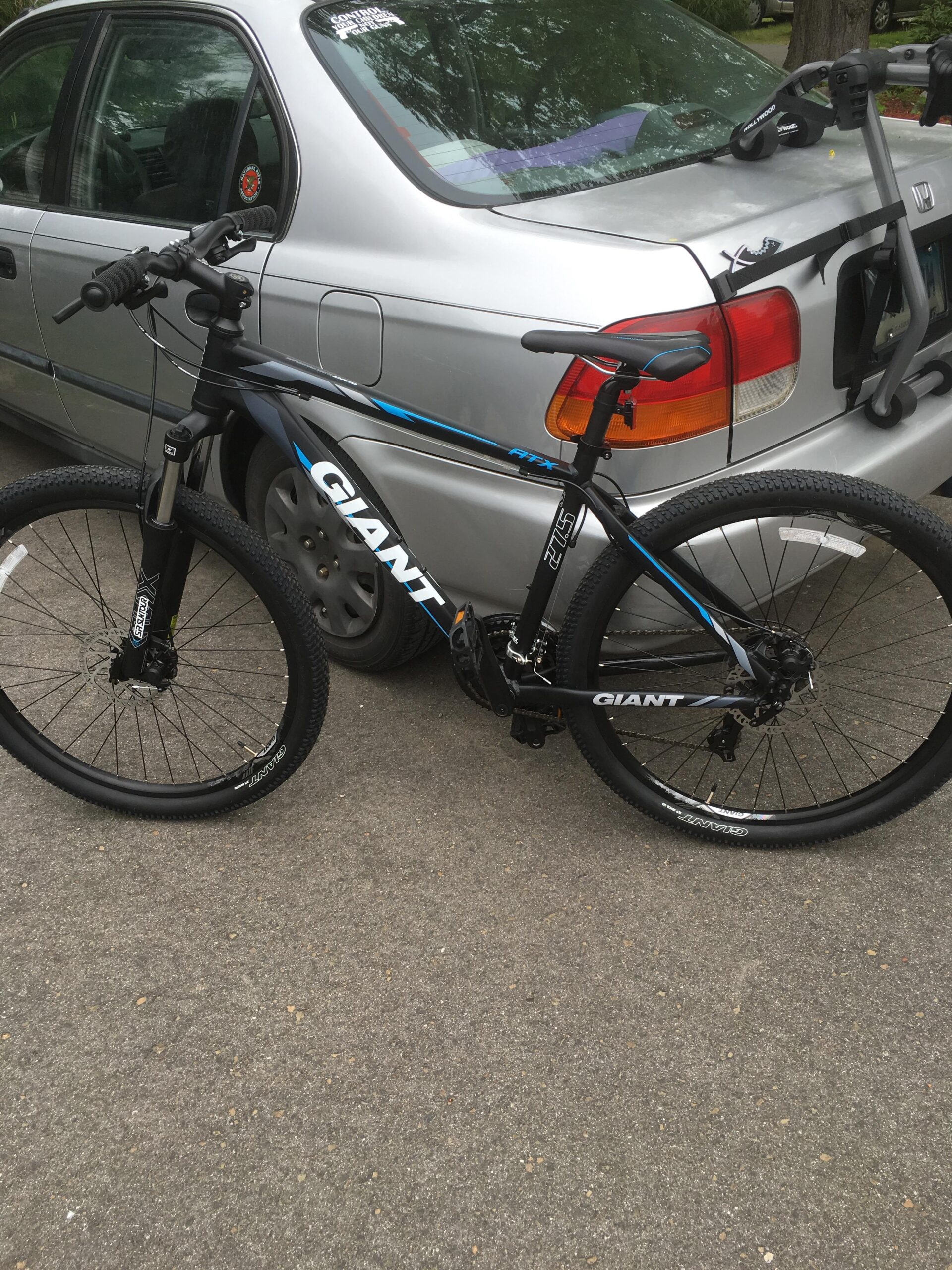 Giant ATX 27.5 2: A black and blue Giant mountain bike parked next to a silver sedan on a paved surface. The bike features thick tires, a front suspension fork, and ergonomic handlebars. The car is partially visible in the background, showcasing its rear with a bike rack attached.
