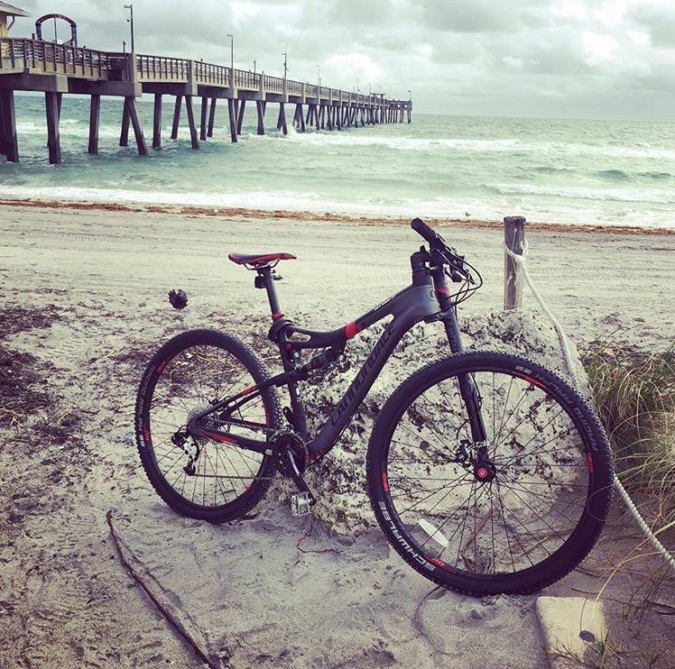 Cannondale Scalpel 29er 3: A mountain bike resting on sandy beach terrain, with a wooden pier extending into the ocean in the background. The sky is overcast, and gentle waves can be seen lapping at the shore. Sparse vegetation is visible nearby.