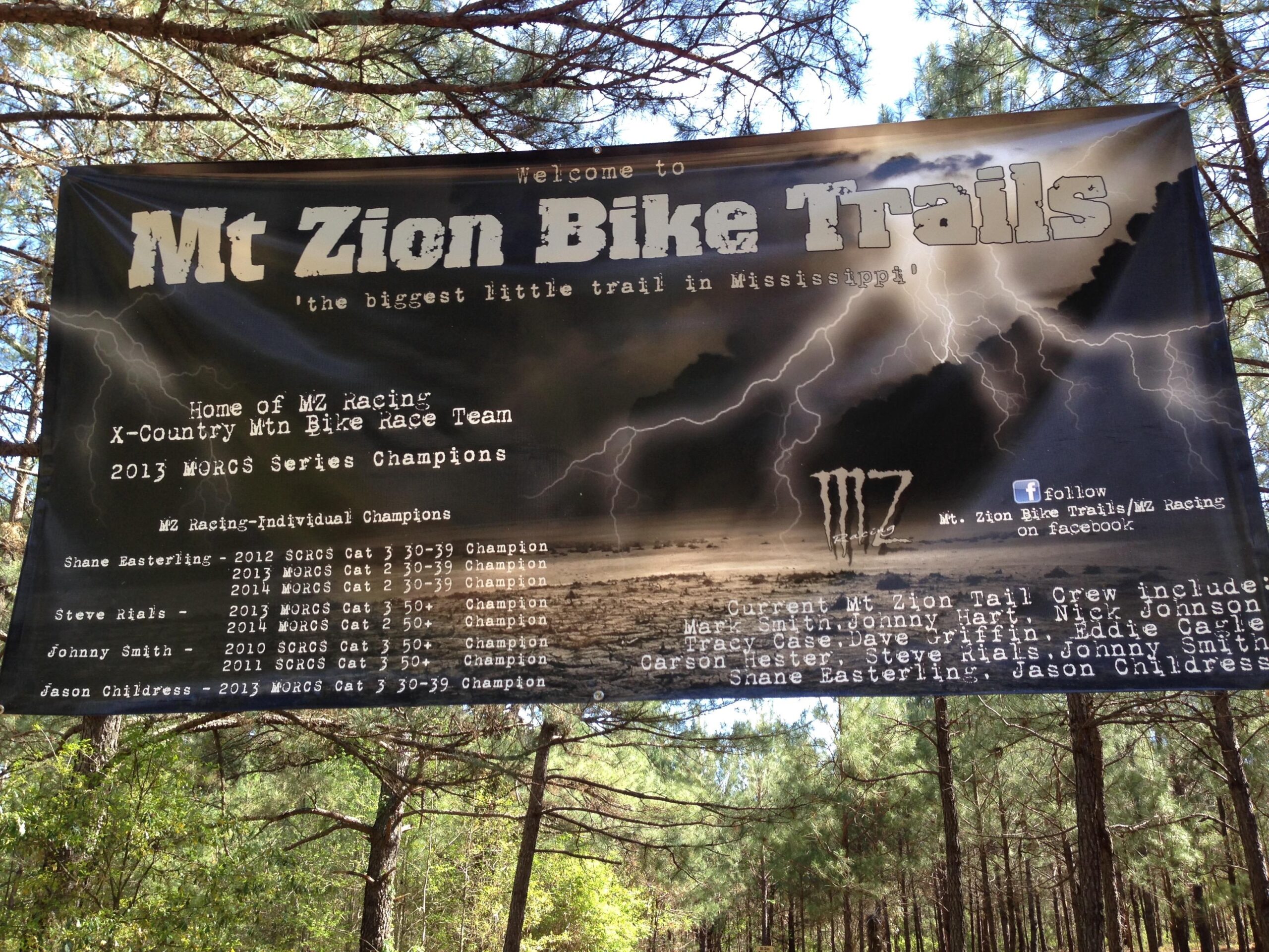 Banner welcoming visitors to Mt. Zion Bike Trails, featuring information about the X-Country Mountain Bike Race Team, the 2013 MORCS Series Champions, and individual champions' names and categories. The background includes a forest scene with trees and a cloudy sky. Mt. Zion Bike Trails mountain bike trail.