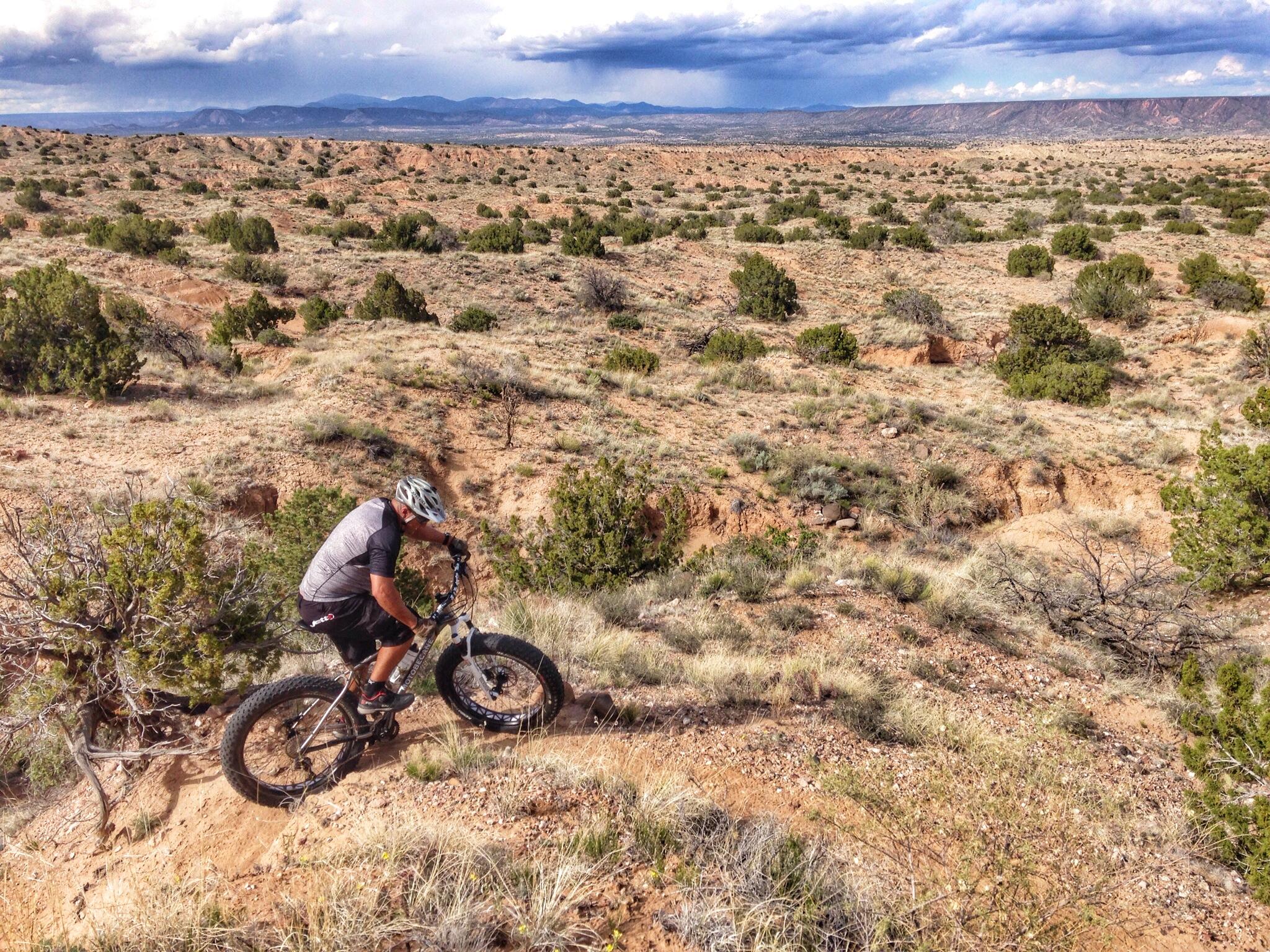 A mountain biker navigates a rugged trail in a desert landscape, surrounded by sparse vegetation and rolling hills. The sky is partly cloudy, with distant mountains visible in the background. The biker is focused on the trail ahead, riding a fat-tire bike on a sunlit path. Mariposa Fat Bike Trails mountain bike trail.
