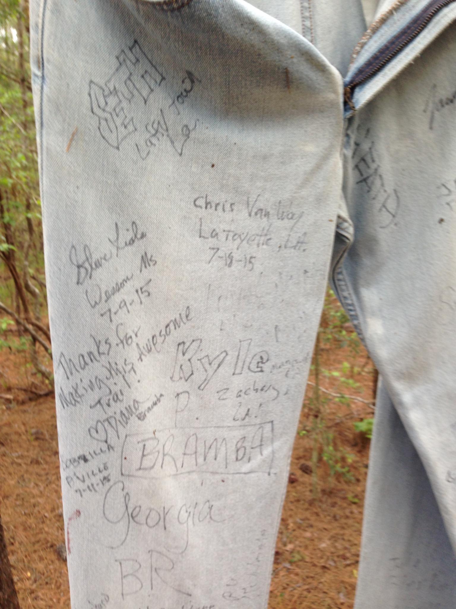 A close-up image of a pair of denim jeans hanging in a wooded area, featuring various handwritten messages and signatures in black ink. The jeans display names, dates, and phrases, showcasing a collection of personal inscriptions from different individuals. Mt. Zion Bike Trails mountain bike trail.