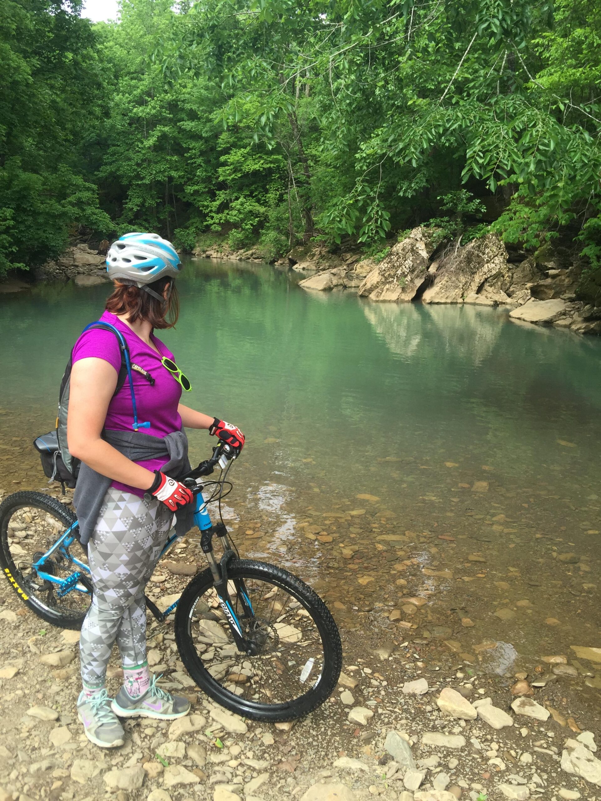 A person wearing a helmet and sunglasses stands beside a clear, turquoise creek, with a mountain bike resting on the ground. They are dressed in a purple shirt and patterned leggings, surrounded by lush green trees and rocky terrain. The tranquil water reflects the greenery, creating a peaceful outdoor scene. Fossil Flats mountain bike trail.