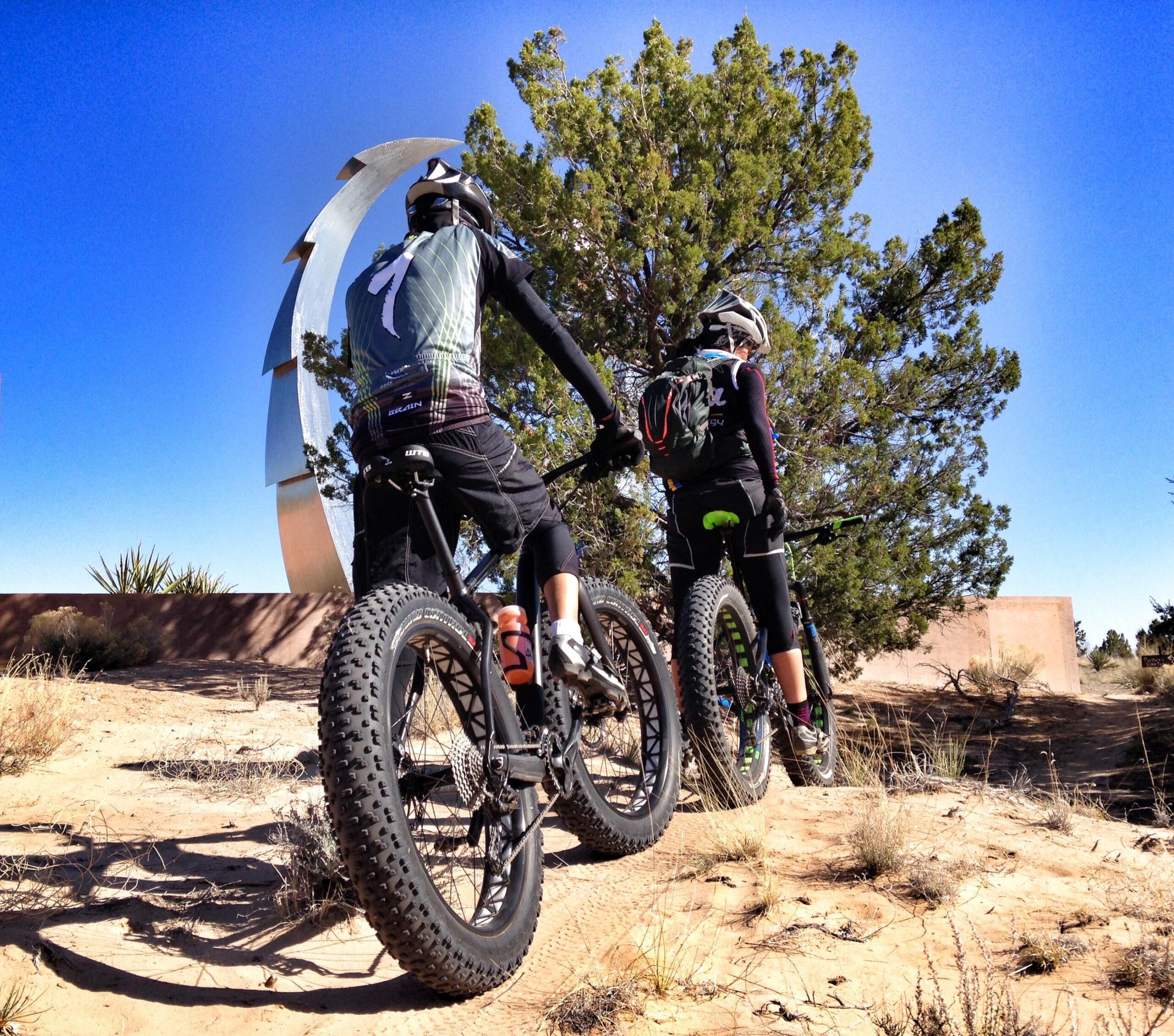 Two mountain bikers standing on a sandy trail, facing a large metal sculpture against a clear blue sky. They are wearing cycling gear and helmets, with one biker’s bike featuring thick tires. Surrounding them are shrubs and bushes typical of a desert landscape. Mariposa Fat Bike Trails mountain bike trail.