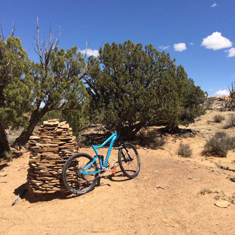 A blue mountain bike resting against a stacked stone formation in a desert landscape, with sparse vegetation and a clear blue sky in the background. High Desert Trail System mountain bike trail.