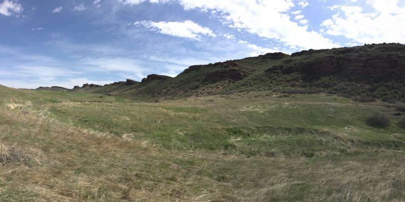 A panoramic view of rolling green hills with rocky outcrops under a partly cloudy blue sky. The landscape features tall grass in the foreground and rugged terrain in the background, creating a serene and natural setting. Devil's Backbone mountain bike trail.