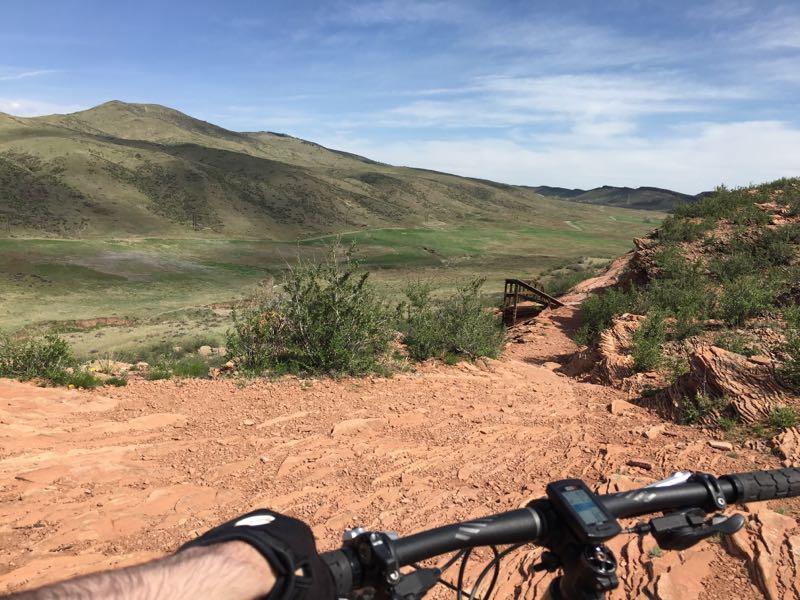 A view from a mountain bike handlebar overlooking a sandy trail leading downhill towards a green valley. In the background, rolling hills are visible under a clear blue sky, with scattered patches of vegetation. A wooden bridge can be seen crossing a small pathway in the distance. Devil's Backbone mountain bike trail.