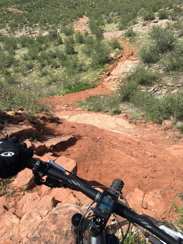 Image showing the handlebar and front wheel of a mountain bike positioned on a rocky, red dirt trail. The trail slopes downward, surrounded by green vegetation and small shrubs, indicating a mountainous terrain in bright sunlight. A bike computer is visible on the handlebars, displaying statistics. Devil's Backbone mountain bike trail.
