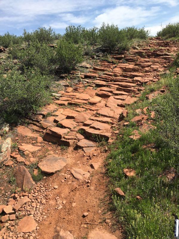 A hiking trail made of uneven red stone slabs, surrounded by green shrubs and grass, leading uphill under a partly cloudy sky. Devil's Backbone mountain bike trail.