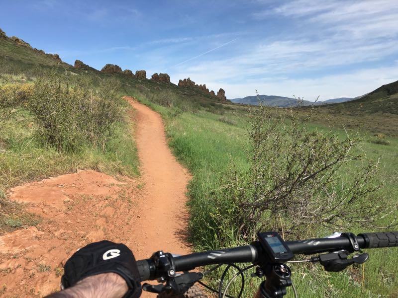 Alt text: A close-up view of a mountain bike's handlebars on a dirt trail surrounded by green grass and shrubs, with rocky outcrops in the background and blue sky above. Devil's Backbone mountain bike trail.