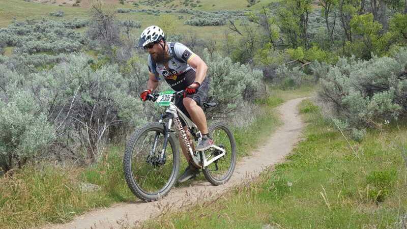 A cyclist is riding a mountain bike along a dirt trail surrounded by shrubs and greenery. The rider, wearing a helmet and cycling gear, is focused on navigating the terrain as he ascends a slight incline. The background features rolling hills and lush vegetation, representing a scenic outdoor setting for mountain biking. Spring Valley mountain bike trail.