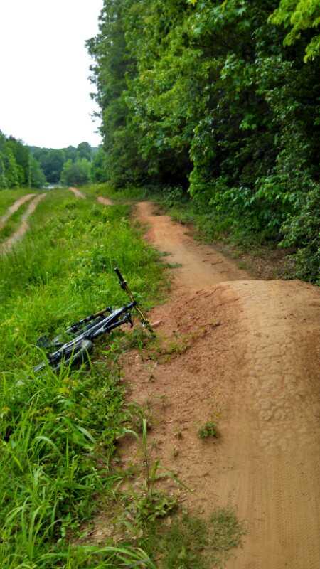 A narrow dirt path surrounded by greenery, with a partially visible mountain bike resting on its side in the foreground. The path leads into a wooded area, and the scene is bright and inviting, suggesting an outdoor recreational space. USNWC mountain bike trail.