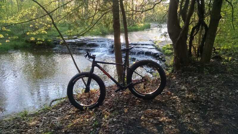 A mountain bike with thick tires is resting on the shore of a serene stream surrounded by lush greenery and trees. In the background, a small waterfall cascades over rocks into the water. Sunlight filters through the foliage, creating a peaceful outdoor scene. Baird Creek mountain bike trail.