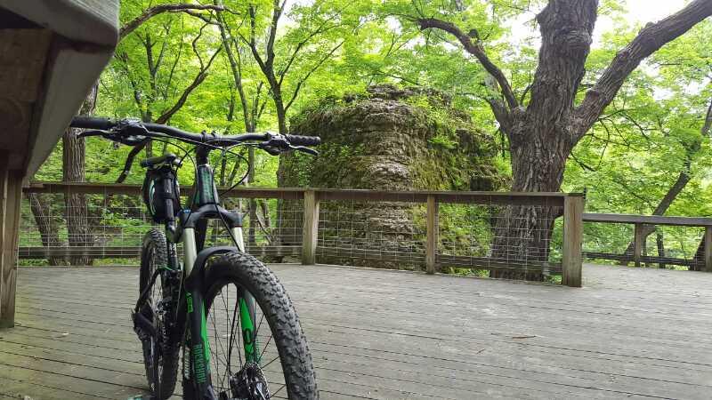 A mountain bike parked on a wooden deck surrounded by lush green trees and foliage, with a moss-covered rock formation visible in the background. Rock Cut State Park mountain bike trail.