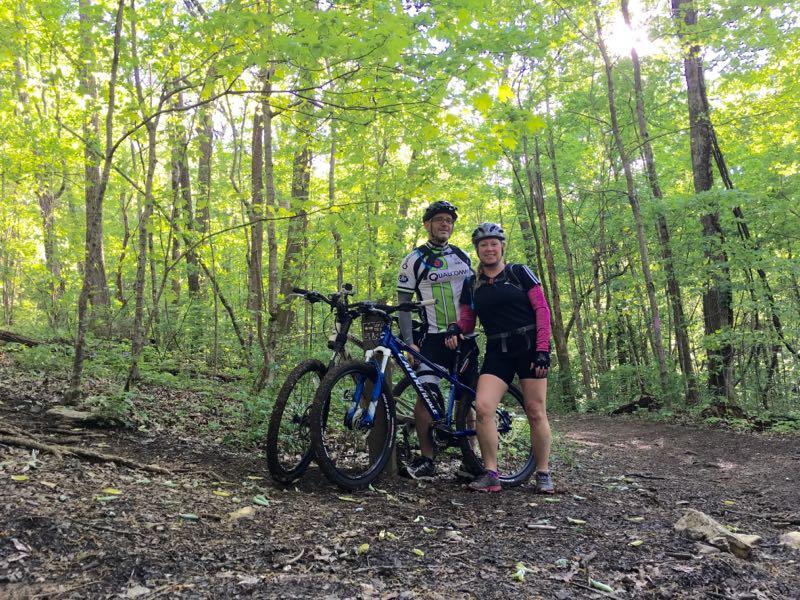 Two cyclists stand beside their mountain bikes on a dirt path surrounded by lush green trees in a forest. The man is wearing a white and black cycling jersey, while the woman is dressed in a black and pink cycling outfit. Sunlight filters through the leaves, creating a bright, natural backdrop. Monte Sano State Park &amp; Land Trust mountain bike trail.
