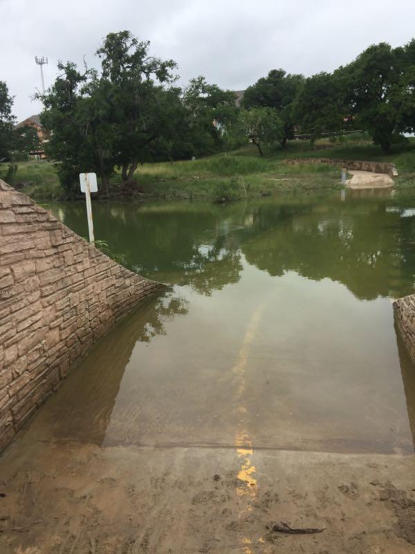 A flooded area showing a submerged road leading into murky water, with a stone edge on the left and trees in the background under a cloudy sky. A white sign is partially visible in the scene. Leon Creek mountain bike trail.
