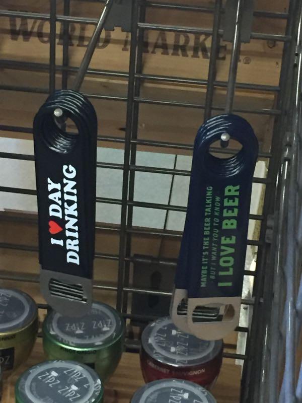 Two colorful drink koozies are displayed on a rack. The koozie on the left reads "I ♥ DAY DRINKING" in white text, while the one on the right says "I LOVE BEER" in green text. Below the right koozie, there is an additional phrase: "Maybe it's the beer talking, but I want you to know." In the background, various cans of beverages can be seen at the bottom. San Tan mountain bike trail.