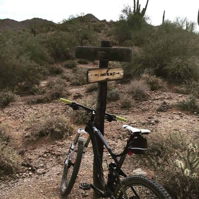 A mountain bike is parked next to a wooden trail sign in a desert landscape, surrounded by sparse vegetation and rocky terrain. The sign indicates various directions for hiking and biking trails, with distant mountains visible in the background under a cloudy sky. Hawes Loop mountain bike trail.