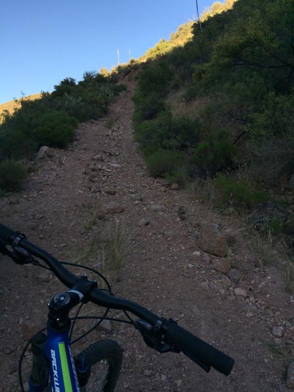 A view of a rugged mountain biking trail leading uphill, with a close-up of a bike's handlebars and front wheel in the foreground. The trail is rocky and surrounded by sparse vegetation and hills under a clear blue sky. Cottonwood Trail #120 mountain bike trail.