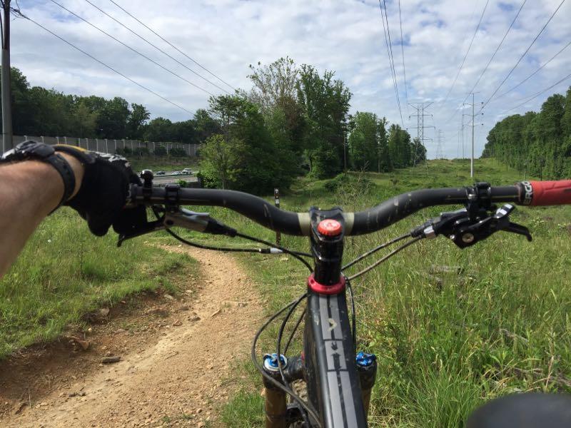 Close-up view of a mountain bike handlebar with a hand gripping the grip. The bike is positioned on a dirt trail surrounded by lush greenery and trees. In the background, power lines run parallel to the trail, and a road can be seen off to the side. The sky is partly cloudy, indicating a pleasant day for riding. Wakefield mountain bike trail.